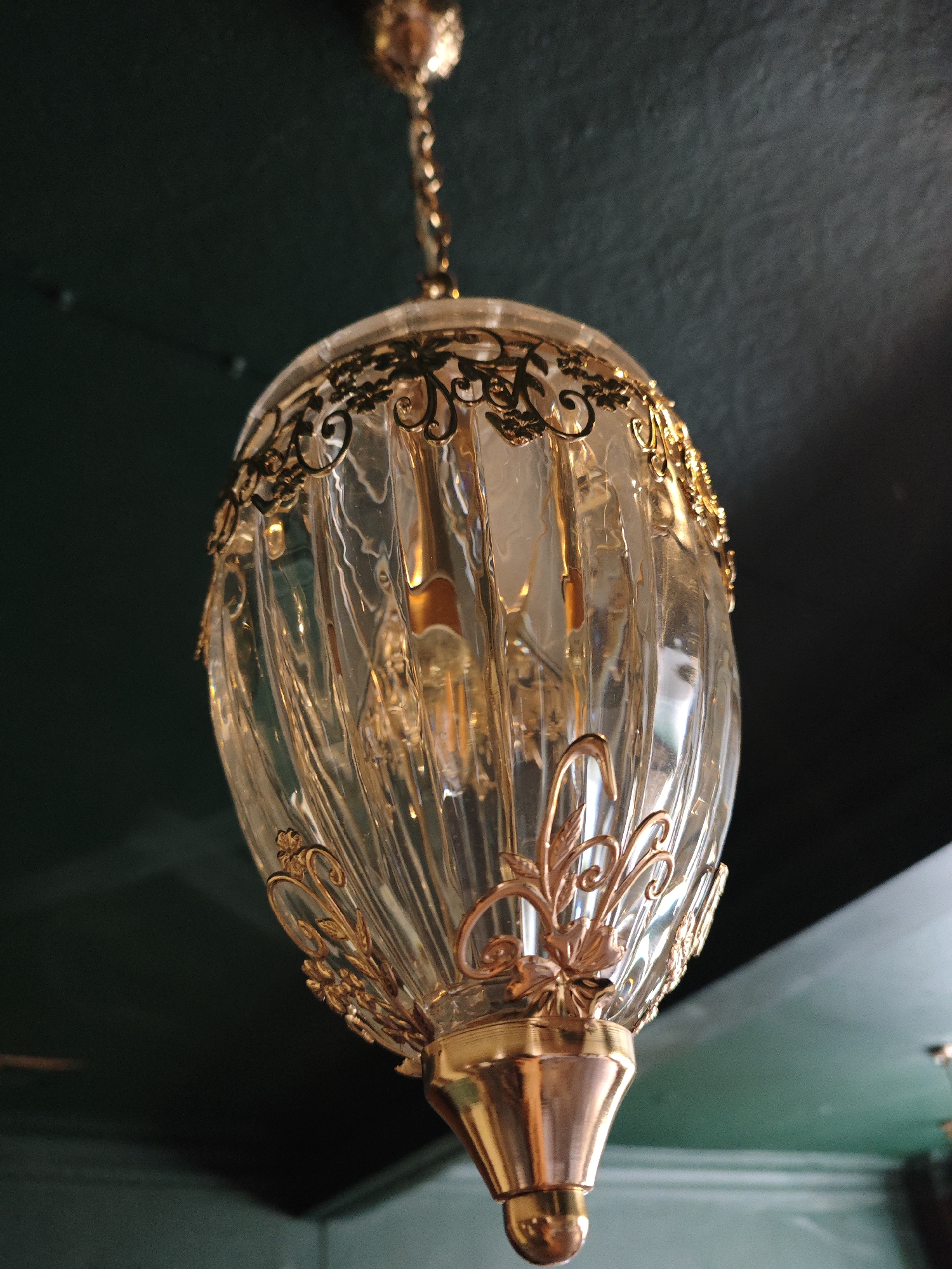 This media file depicts a close-up, low-angle shot of an ornate, clear glass pendant light fixture. The fixture is suspended from the ceiling by a gold-colored chain. The glass shade has a ribbed, elongated oval shape and is adorned with intricate gold metal scrollwork and floral embellishments around the top and bottom. Inside the glass shade, a light bulb is visible, emitting a warm, golden glow that reflects off the glass and metal elements. The ceiling is painted a dark, muted green. The lighting suggests it could be daytime, but the indoor setting and artificial light make it difficult to determine. No people or specific location cues are visible, and there is no visible text. The overall impression is one of elegant, possibly vintage, interior decor.