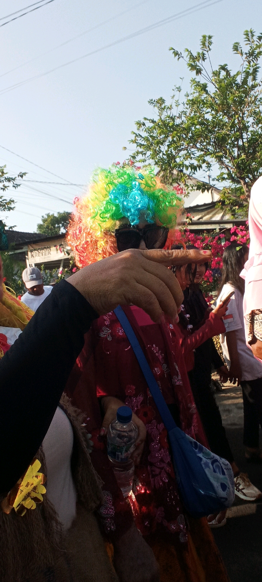 This is a close-up, vertical, outdoor photograph taken in Gempolklutuk, Indonesia, depicting a person in a rainbow-colored wig and dark sunglasses, holding a bottle of water and pointing. The individual is wearing a red, sequined garment. A blue bag with a patterned design hangs from their shoulder. In the background, a sign with the text "REGION KRETER" is visible. Other people are present, including a person wearing a white shirt and a grey cap, and several women in the middle ground. Pink flowers and green trees form part of the environment. The lighting suggests it is daytime, likely sunny, casting shadows. The foreground is dominated by the pointing hand and the colorful wig.