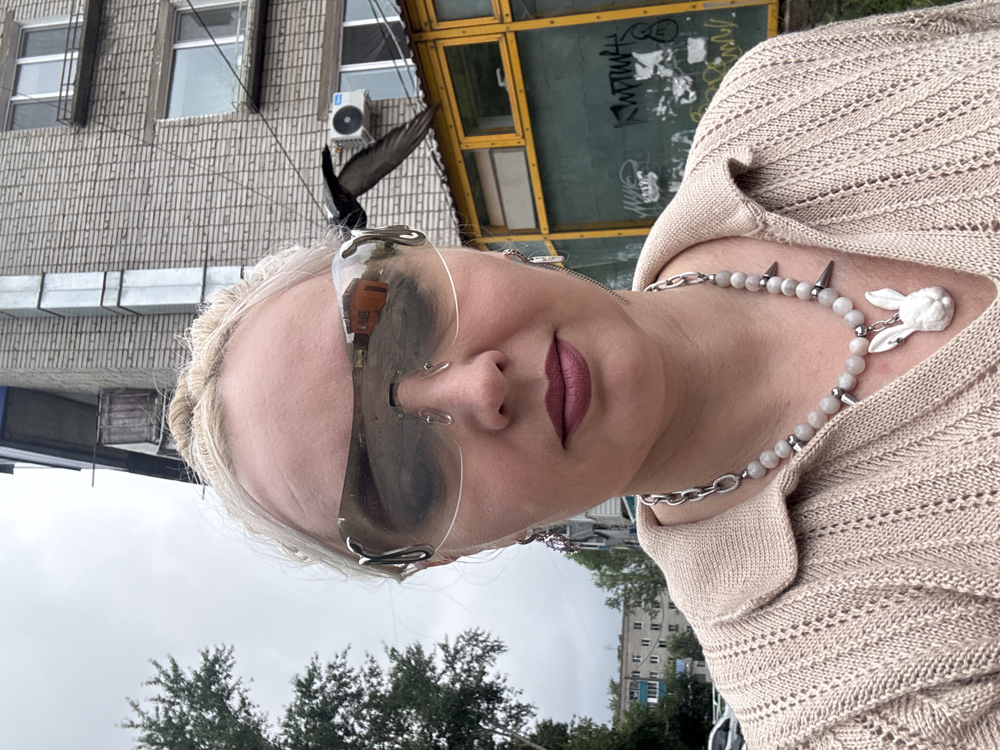 A woman with blonde hair pulled back and wearing large, avant-garde sunglasses takes a selfie outdoors in front of a brick building. She has on a light-colored knitted top with a collared neckline and a necklace with grey beads, silver hardware, and a white rabbit pendant. She is also wearing dangling earrings. The building appears to be an apartment complex with balconies and an air conditioning unit visible. There is a yellow-framed structure with graffiti on the lower portion of the building. Trees and other buildings are visible in the background, suggesting an urban environment. The sky is overcast. The scene is located in Komsomolsk-on-Amur, Russia.