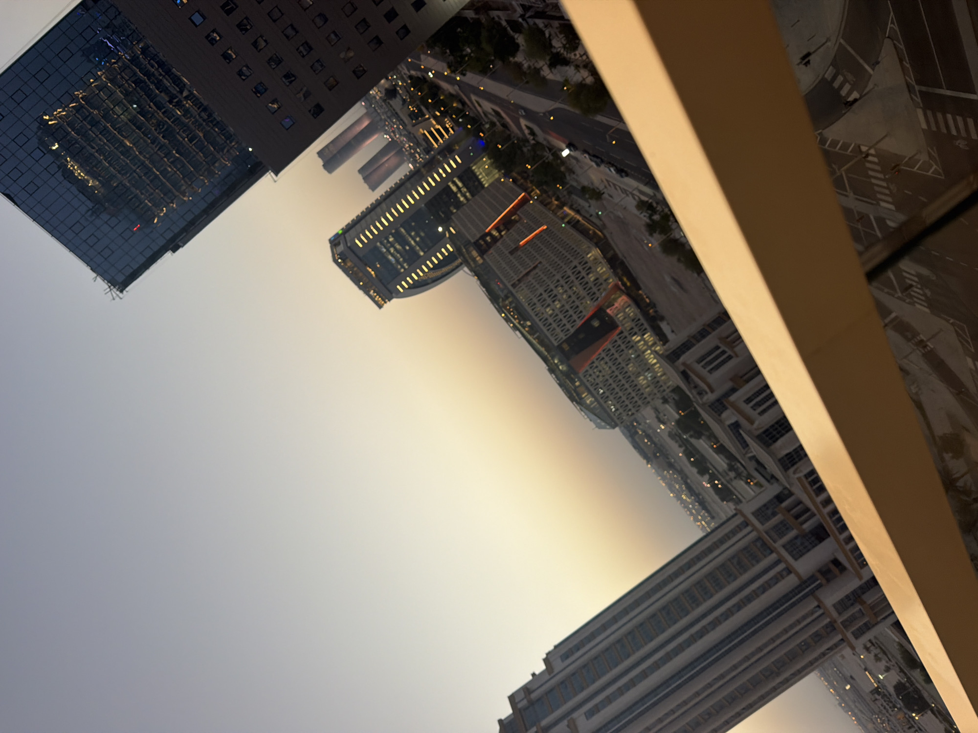 The image shows a city skyline at dusk or dawn. The sky is a gradient of light gray to pale yellow. In the foreground, a beige railing runs diagonally across the bottom of the frame. Beyond the railing, several tall buildings are visible, some with illuminated windows, suggesting it is nighttime or twilight. The buildings are modern architectural structures with glass facades and sharp angles. One prominent building on the right has a dark, reflective glass top that mirrors another building. The perspective is angled, giving a sense of looking down from a height onto the urban landscape. Streets and lights are visible in the distance, indicating a densely populated area. There are no discernible people or active interactions depicted in the scene. The overall impression is one of a contemporary city at a transitional time of day.