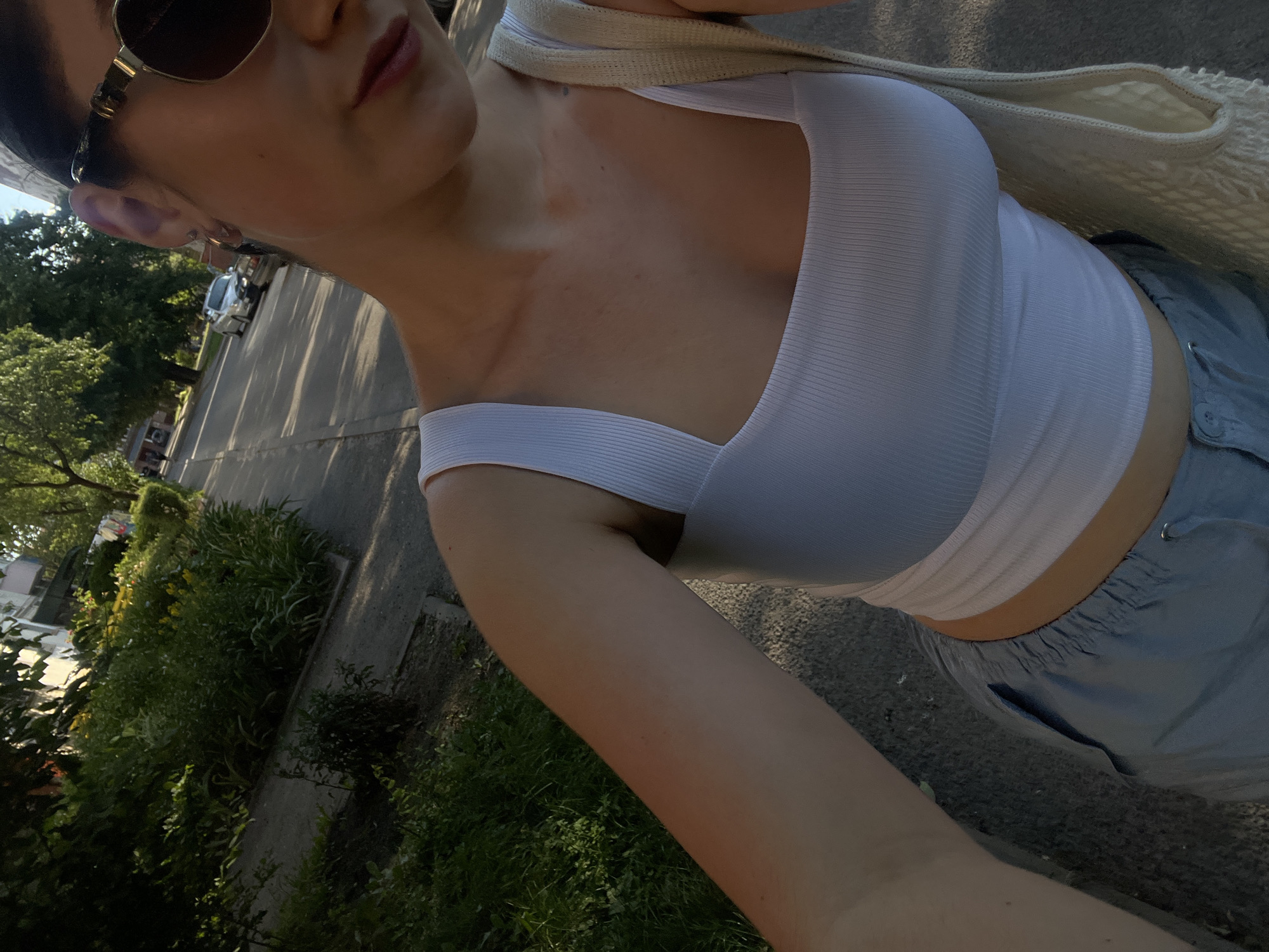 The image shows a person from the chest down, wearing a white ribbed tank top and light blue shorts. The person is outdoors, with a tree-lined street and sidewalk visible in the background. Sunlight casts long shadows, suggesting it is either early morning or late afternoon. The person is holding a woven bag over their shoulder. The overall tone of the image is casual and relaxed.