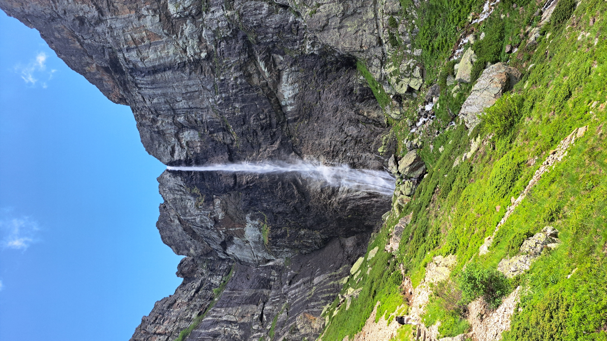 A person is hiking on a rocky trail next to a steep, grassy slope leading up to a towering waterfall cascading down a sheer, dark rock face. The waterfall is the central focus of the image, with water spraying outwards near its base. The surrounding cliffs are rugged and imposing, with streaks of lighter rock and patches of green vegetation clinging to their surfaces. The sky above is a clear, bright blue with a few wispy clouds. The terrain is mountainous and wild, suggesting a remote natural landscape.