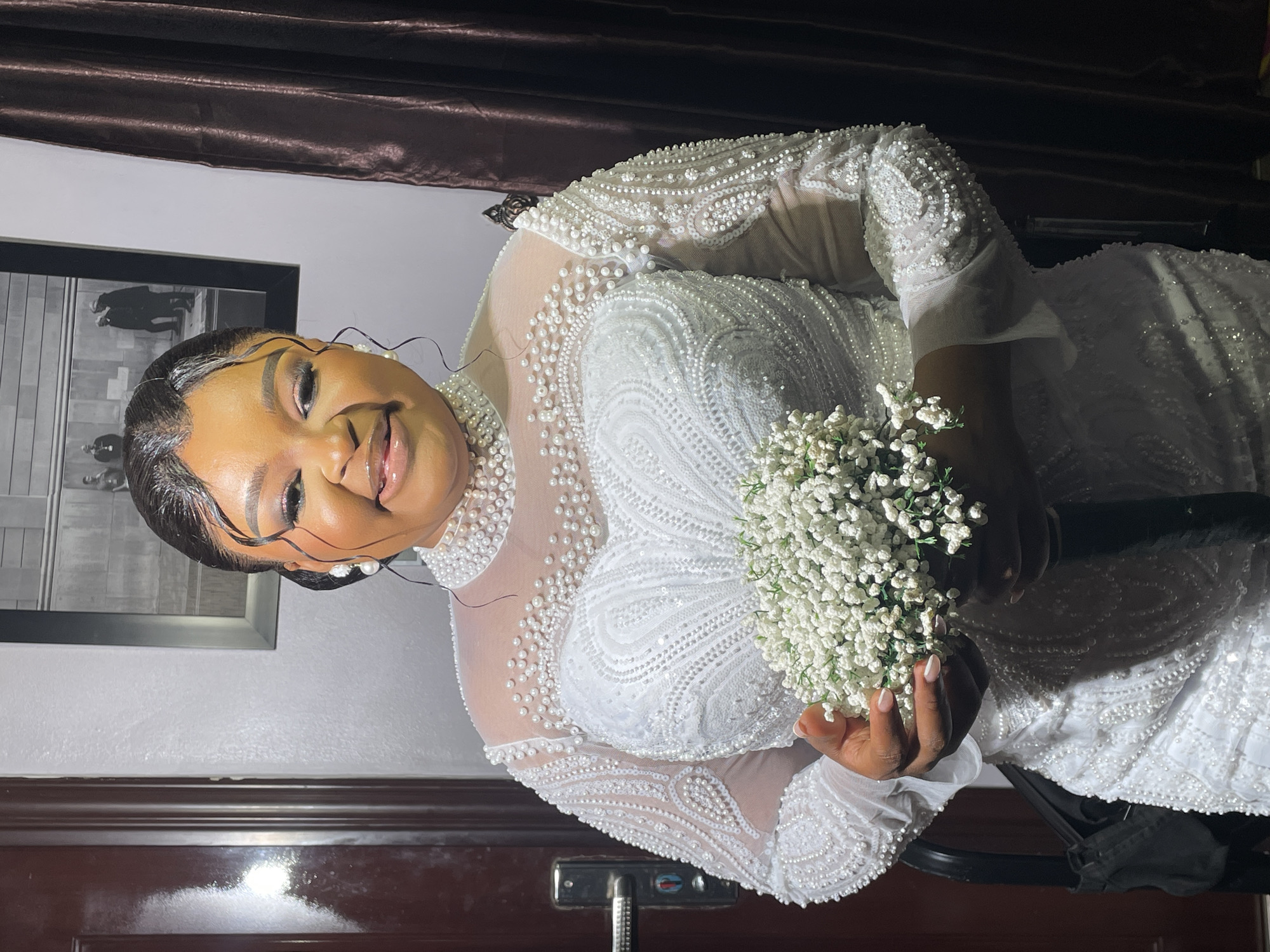 A dark-skinned woman in a white, pearl-encrusted wedding dress holds a bouquet of white baby's breath flowers. She smiles and looks towards the viewer. She has her hair styled in an updo with strands framing her face. In the background, there is a framed picture on the wall, a brown curtain, and a dark brown wooden door. The photo was likely taken indoors, but the exact location in Bar Jirgi Summa, Nigeria is not discernable from the image.