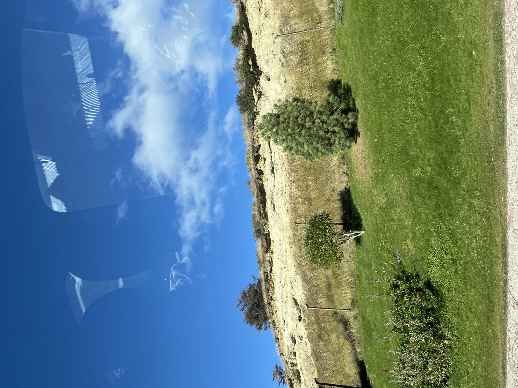 The image shows a view from a vehicle with a sky of blue and white clouds. A cliff with trees and grasses is visible to the right. The location and the city are not discernible from the image.