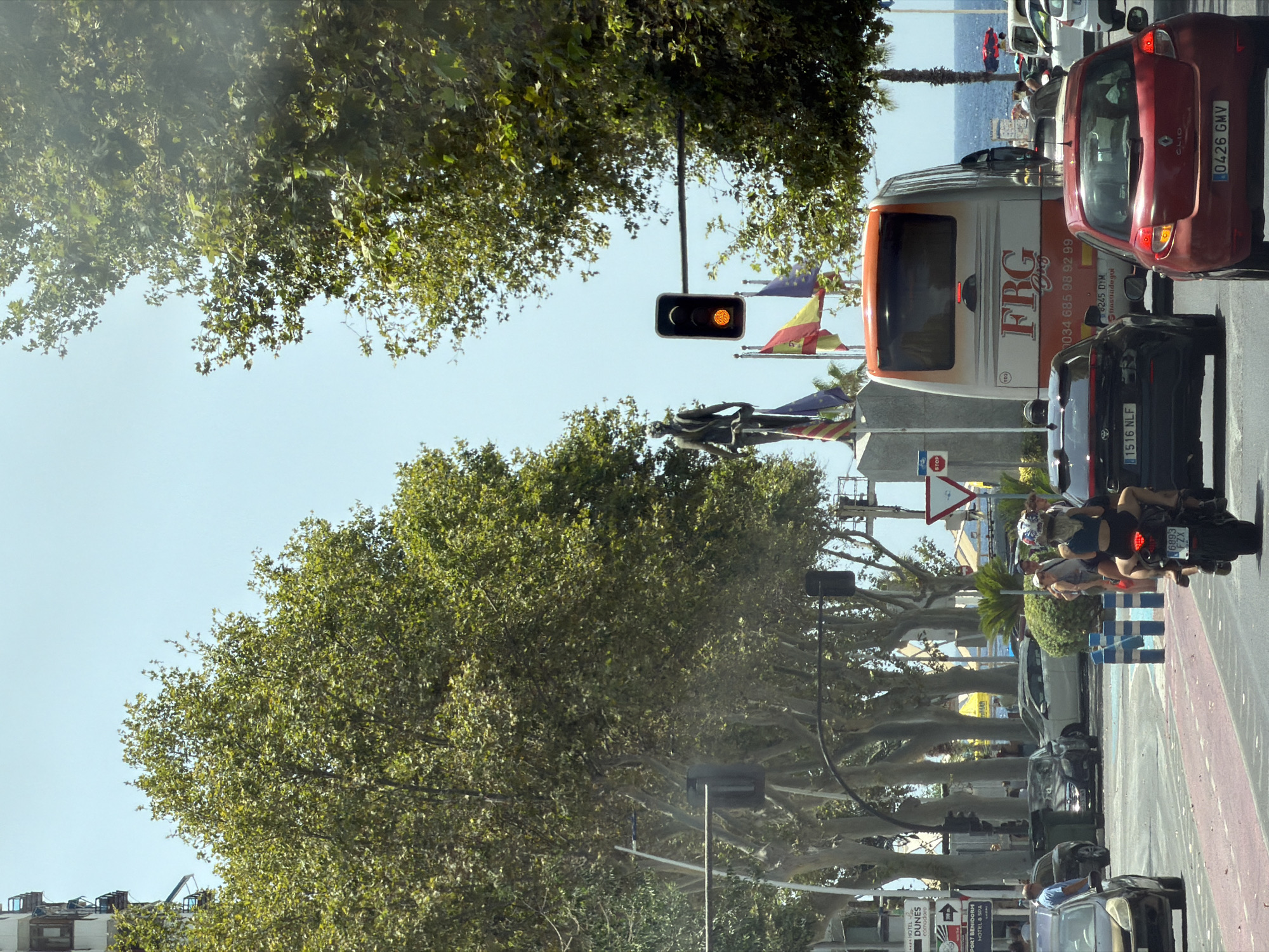The image depicts a street scene in Benidorm, Spain, on a sunny day. A statue of a figure standing on a pedestal is visible on the left, flanked by flags of Spain and the European Union. A traffic light with an amber signal is illuminated. A large, orange and white bus with "FRG" and a phone number printed on it is in the center of the road, behind a black car with license plate "1516 NLF." To the right of the bus, a red Renault Clio with license plate "0426 GMV" is also in traffic. A motorcycle with two occupants, a driver and a passenger, is in the foreground, heading away from the viewer. The background shows a row of trees lining the street, with buildings and the sea visible in the distance. Several other vehicles and pedestrians are present, suggesting a busy urban environment.