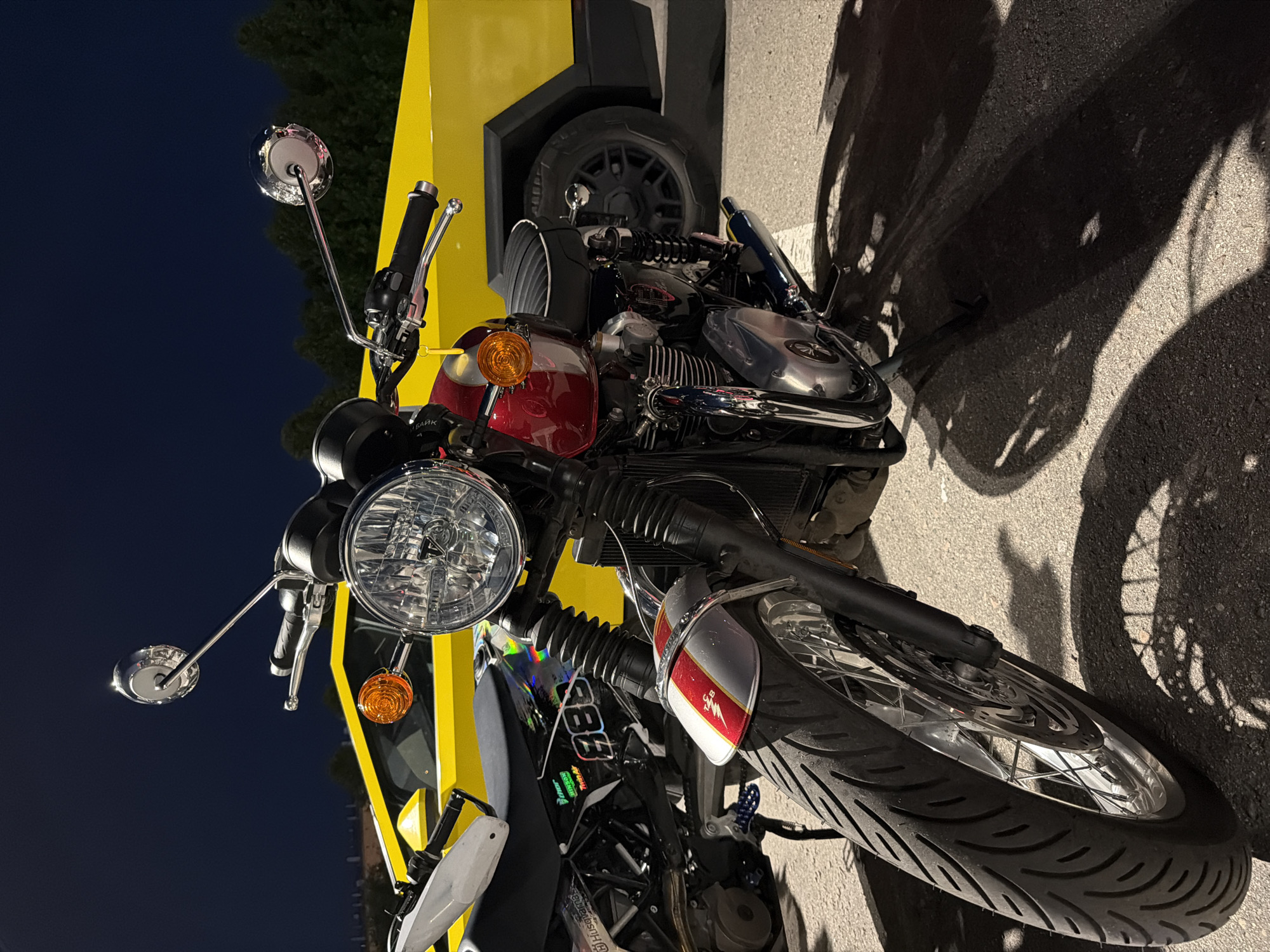 An image captures a vintage-style motorcycle parked at night, illuminated by an artificial light source that casts long, distinct shadows on the asphalt ground. The motorcycle is presented in a dynamic, angled composition, with the original image rotated 90 degrees counter-clockwise, making the ground appear vertical.
The main subject is a classic-looking motorcycle featuring a prominent round chrome headlight, chrome mirrors, and orange turn signals. Its fuel tank is a glossy deep red, adorned with chrome accents. Below the handlebars, on the tank, the partially visible Russian word "БАЙК" (BAYK, meaning "bike") is present. Further back, a black panel beneath the tank displays the number "983" and additional smaller text, including "Husqvarna". The motorcycle has spoked wheels, and its front fender is silver-grey with a red and white stripe pattern, featuring a small "M&B" logo and a stylized winged emblem.
Behind the motorcycle, a bright yellow structure dominates the background, possibly part of a building or a large sign. Nestled between the motorcycle and this yellow structure, a dark grey car tire and wheel are visible, suggesting another vehicle or a display. The background sky is a dark, clear blue, indicating nighttime. Shadows of the motorcycle's components, including its wheels and handlebars, stretch across the textured asphalt. No people are visible, and the motorcycle appears to be stationary. The overall scene suggests an urban or roadside setting in Moscow, Russia, during the evening.