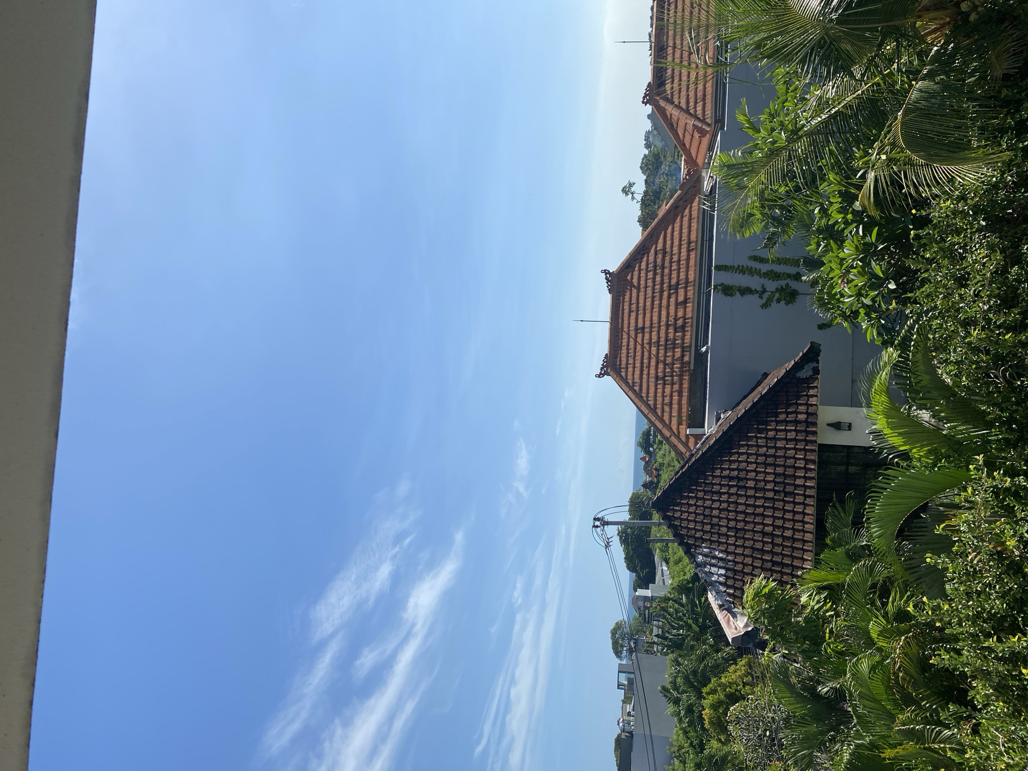 The image shows a view in North Kuta, Indonesia, featuring several buildings with traditional tiled roofs. Lush green vegetation, including palm trees and various shrubs, surrounds the buildings. Overhead power lines and poles are visible. The sky is blue with wispy clouds. The image appears to have been taken from an elevated position, possibly a balcony or higher floor of a building.
