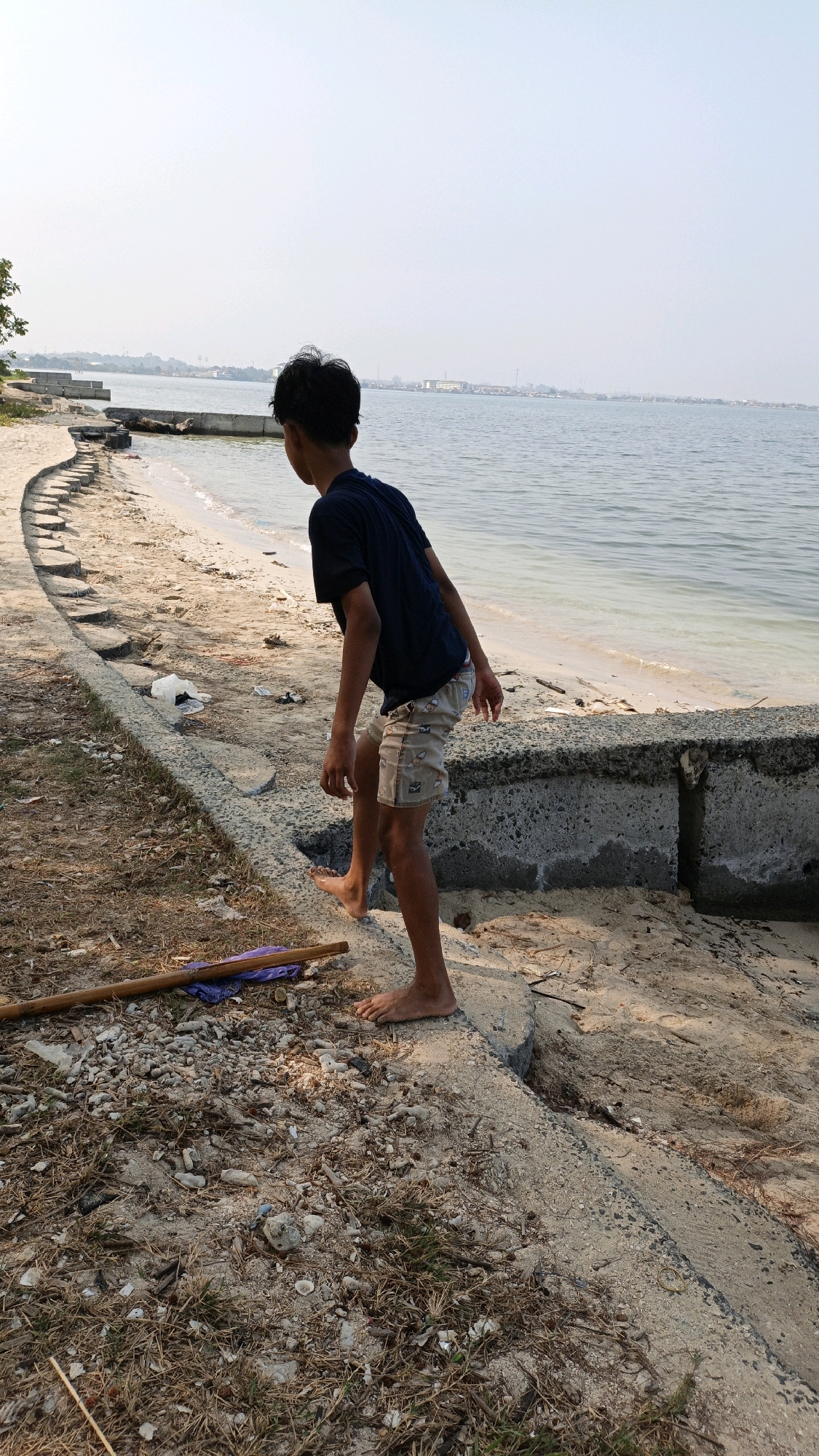 A young boy, viewed from the side, stands on a sandy shore in Cikal, Indonesia. He is wearing a dark blue t-shirt and patterned shorts. His right leg is bent, and his right foot is lifted as if he is about to step down onto the sand. The boy is facing away from the camera, looking towards the water.
The shore is a mix of sand, dry grass, and some debris. A concrete structure runs along the edge of the shore, with some sections broken or eroded. The water is calm and appears to be a bay or sheltered area, with land visible in the distance. The sky is overcast and bright, suggesting it is daytime. A wooden stick and a purple cloth are lying on the ground near the boy's feet.