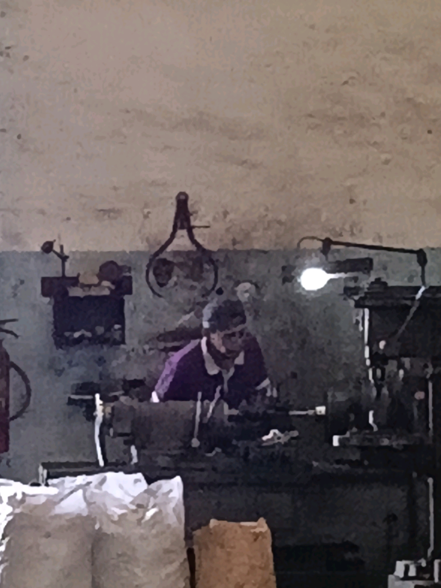 The image depicts an indoor scene, possibly a workshop or factory, in Belimbing, Indonesia. A person wearing a purple shirt is seated at a workbench, appearing to be engaged in some form of manual labor, possibly operating machinery. The environment is somewhat cluttered with various objects, including what appear to be tools, equipment, and bags of material in the foreground. A light fixture is visible on the wall, illuminating the workspace. The overall impression is of an industrial or artisanal setting.
