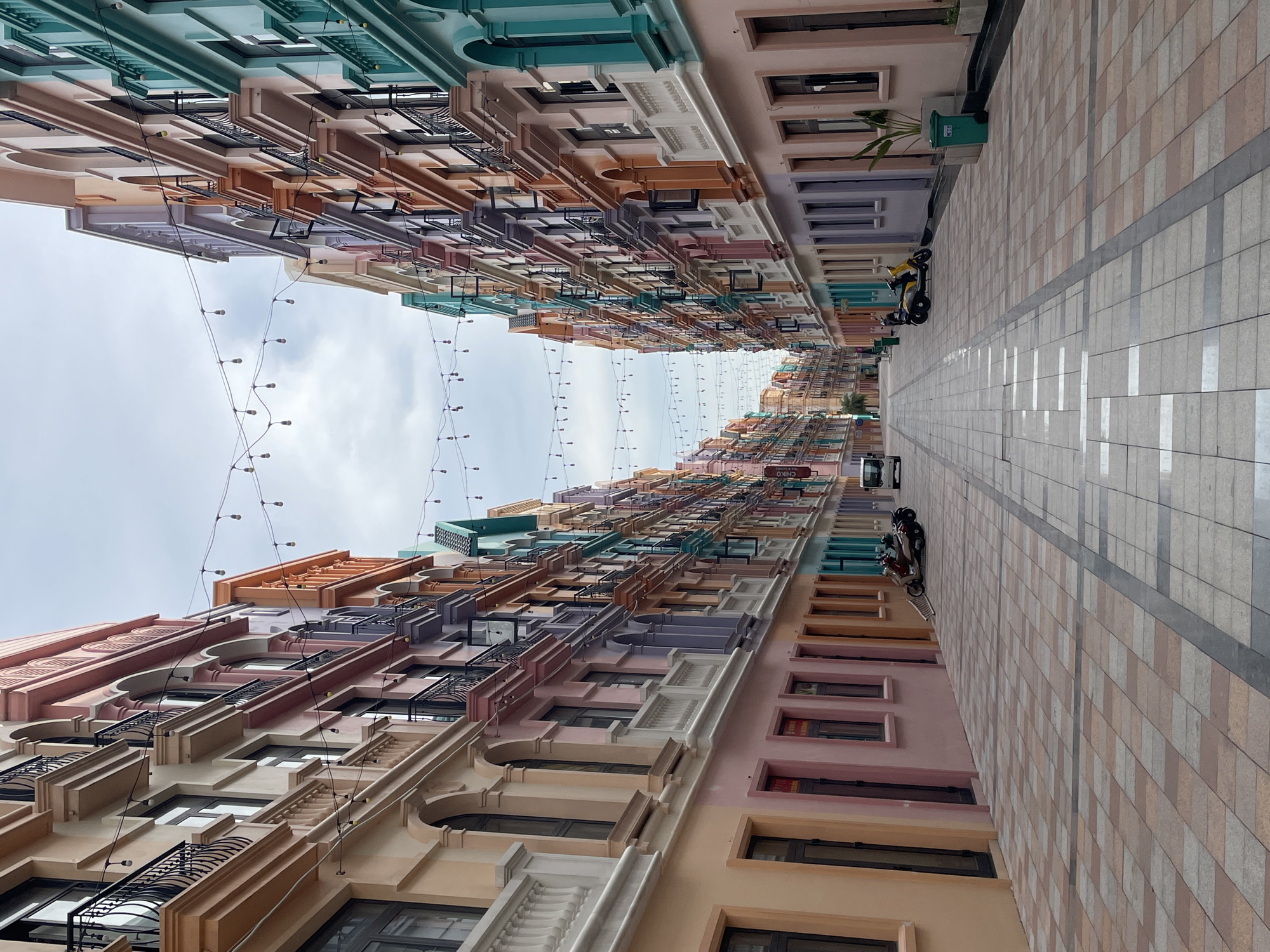 The image shows a street in Nghĩa Trụ Commune, Vietnam, lined with colorful buildings. The buildings are painted in pastel shades of pink, peach, lavender, and teal, with architectural details like balconies, arched windows, and decorative cornices. String lights are hung overhead, illuminating the walkway. The street is paved with patterned tiles, and there are a few motorcycles parked on the side. A white van is visible in the distance. The sky is overcast.