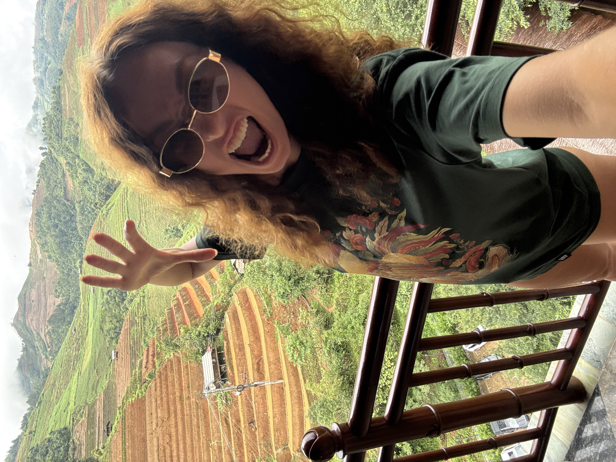 A young woman with long, curly, reddish-brown hair is captured in a dynamic selfie, rotated 90 degrees counter-clockwise. She is positioned on a balcony, with a dark wooden railing partially visible in the foreground. The woman wears round, gold-rimmed sunglasses and a dark green t-shirt adorned with a colorful, possibly floral or avian, design. Her mouth is wide open in an expression of excitement or joy, revealing her teeth. Her left hand is raised towards the camera, fingers spread, adding to the energetic pose.
The background reveals a breathtaking panoramic view of terraced fields and lush green mountains, characteristic of a mountainous region in Púng Luông, Vietnam. The terraces display varying shades of green, indicating cultivated areas, interspersed with reddish-brown earth. Small structures or houses are dotted across the landscape. The sky appears overcast or hazy, with clouds hugging the mountain peaks. The overall scene suggests a vibrant daytime view, full of natural beauty, with the woman conveying a sense of playful enthusiasm during her travels. There is no discernible text visible in the image.