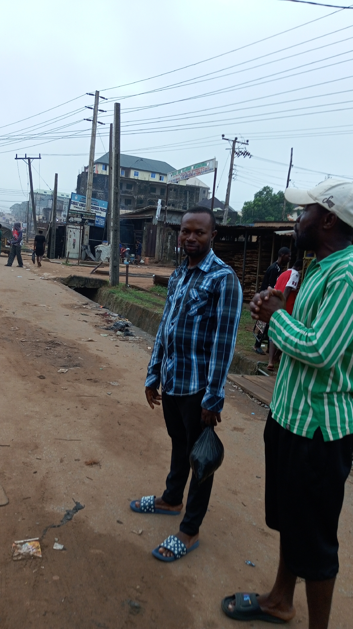 The image shows two men standing on a dirt road in Onitsha, Nigeria. The first man is wearing a blue and black plaid shirt, black pants, and blue and white sandals, holding a black plastic bag. The second man is wearing a green and white striped shirt, black shorts, a white cap, and black sandals. In the background, there are buildings, utility poles, and other people. The sky is overcast. A sign reads "CHRIST ANGELI".