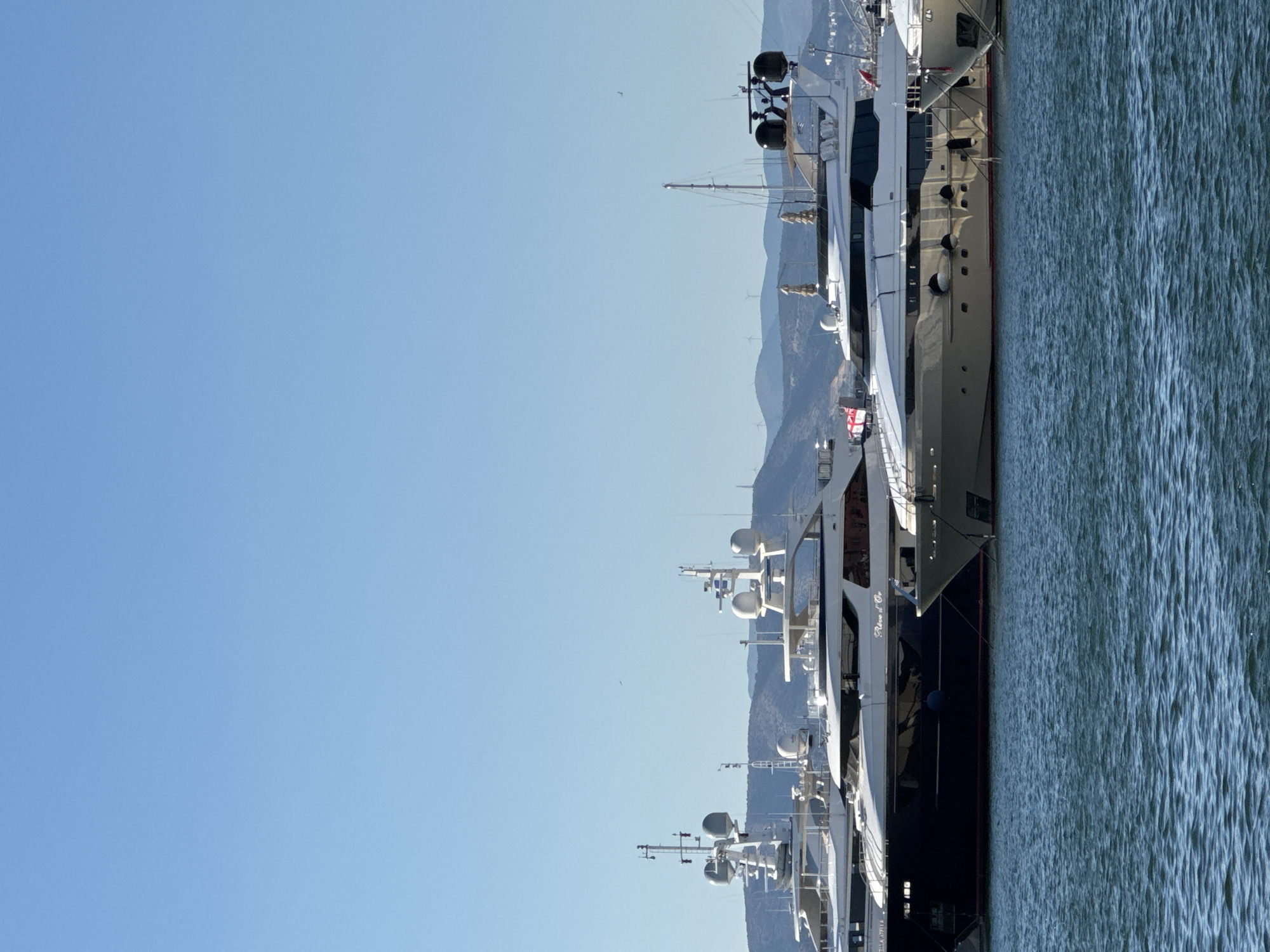 The image shows several large yachts docked in a harbor under a clear blue sky. In the foreground, dark blue and white yachts are visible, with the name "Rêve d'Or" inscribed on one of them. The yachts have multiple decks, radar domes, and what appear to be satellite dishes. In the background, rolling hills with wind turbines can be seen. The water in the foreground is calm with a slight ripple. The scene is indicative of a luxury marina. The image was taken in Moschato, Greece.