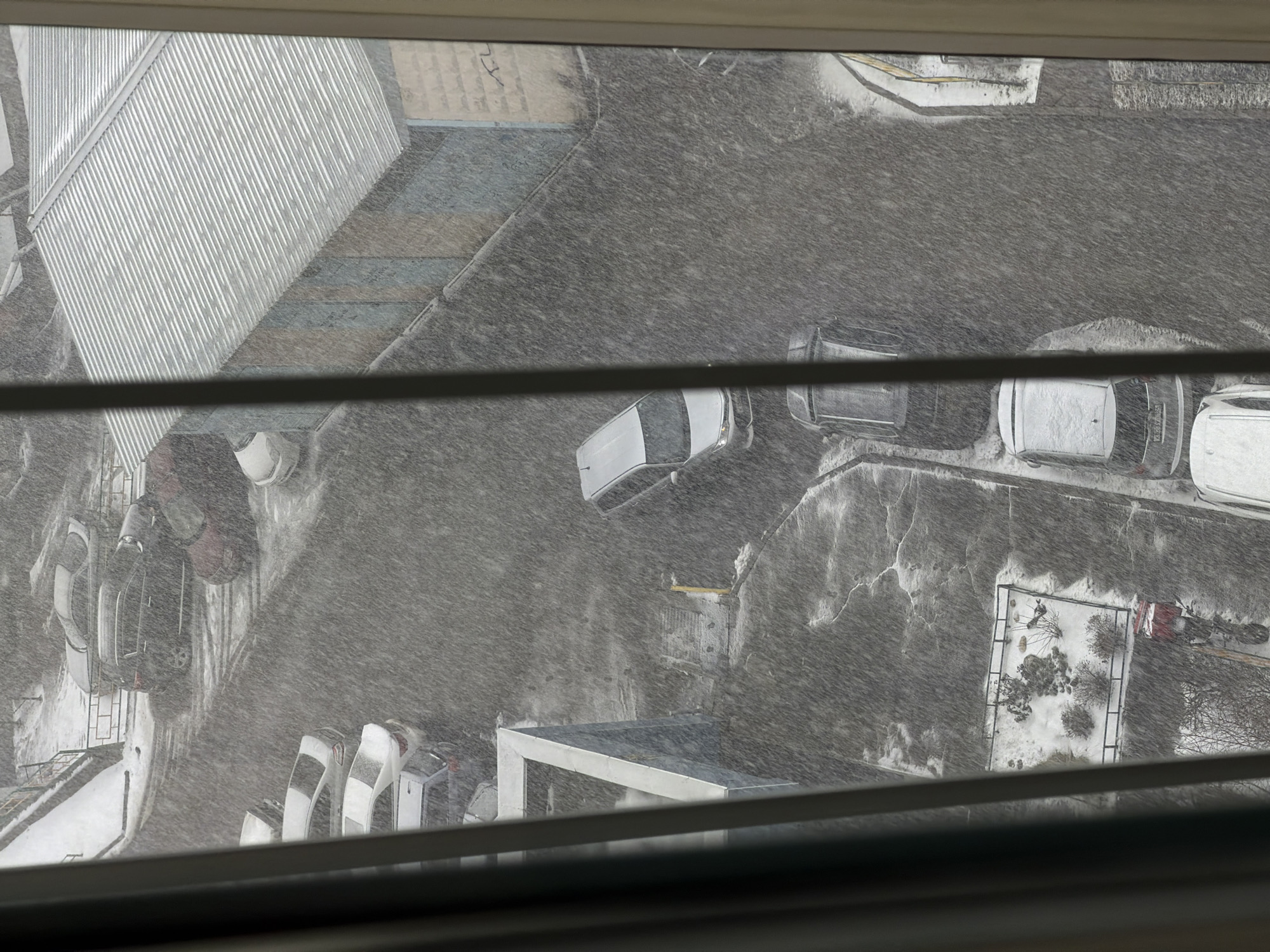 From a high vantage point, likely a window in Krasnoyarsk, Russia, a snowstorm blankets the scene. The view overlooks a parking lot covered in snow, with several cars parked haphazardly, also covered in snow. Some structures are visible, including a corrugated metal roof, concrete walkways, and a small fenced-in area possibly a garden or playground also covered in snow. The snow falls heavily, obscuring details and creating a gray, muted atmosphere. The photograph is taken through window blinds.