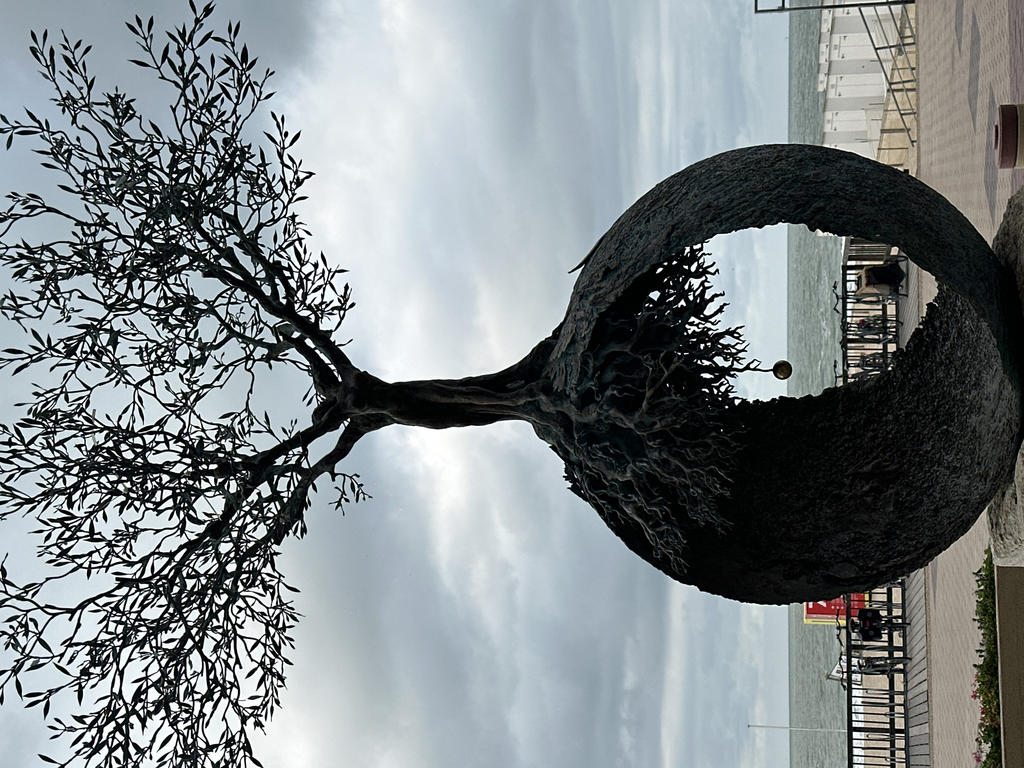 The image shows a striking metal sculpture of a tree with intricate roots emerging from a spherical base. The tree's branches are spread wide, silhouetted against a cloudy sky. The sculpture is positioned outdoors, in front of a beach scene. In the background, the sea is visible under a grey sky, with a beach walkway, railings, beach huts, and parked bicycles. The sculpture is located on the Zoute Avenue in Knokke-Heist, Belgium, a prominent area known for its art installations along the promenade. The atmosphere appears calm, with no individuals actively interacting with the sculpture in the frame.