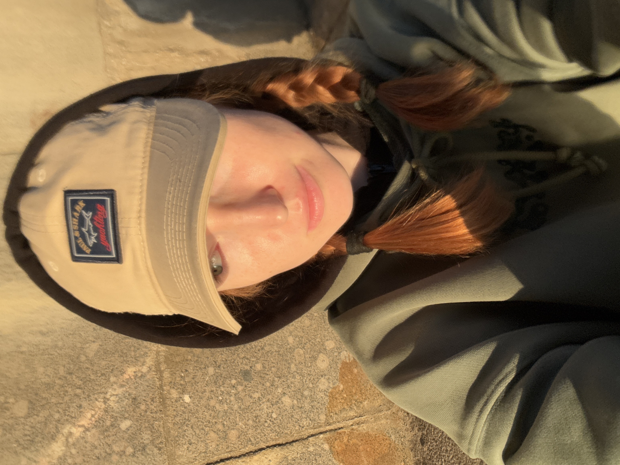 A young woman with auburn hair in braids, wearing a tan baseball cap and a green hooded sweatshirt, is photographed from the chest up, looking to her left. The cap has a patch with the Paul & Shark logo. She is positioned outdoors, with a textured stone wall in the background. The lighting suggests it is either sunrise or sunset, casting long shadows. The woman's face is partially obscured by the cap's brim. The image appears to be a selfie. The background does not provide specific indicators of a location in Saint Petersburg, Russia.