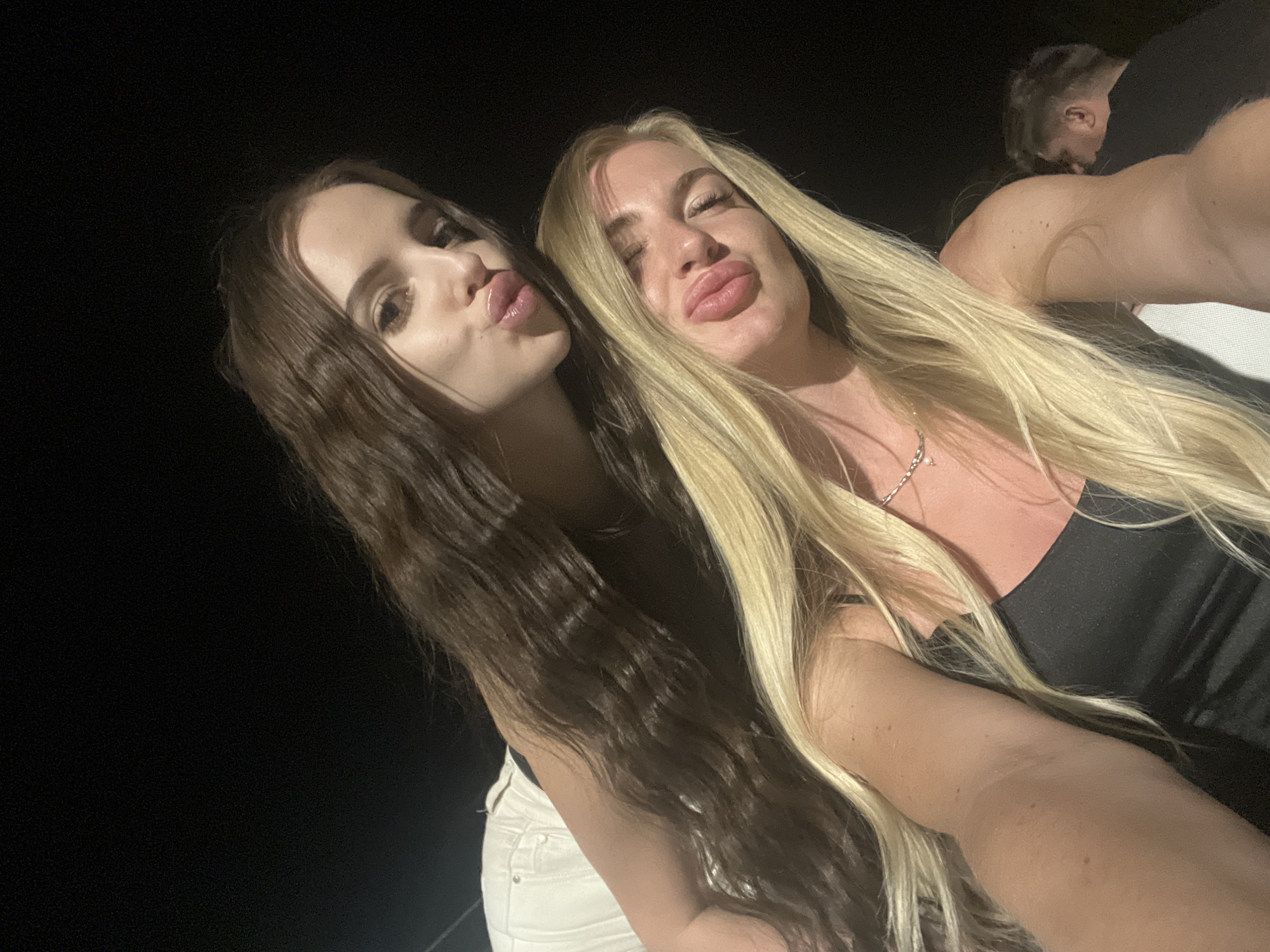 Two women are posing for a selfie. The woman on the left has long, dark brown hair, and the woman on the right has long, blonde hair. Both are making a kissing face. The background is dark, but a third person is partially visible in the upper right corner. The location is not discernable.