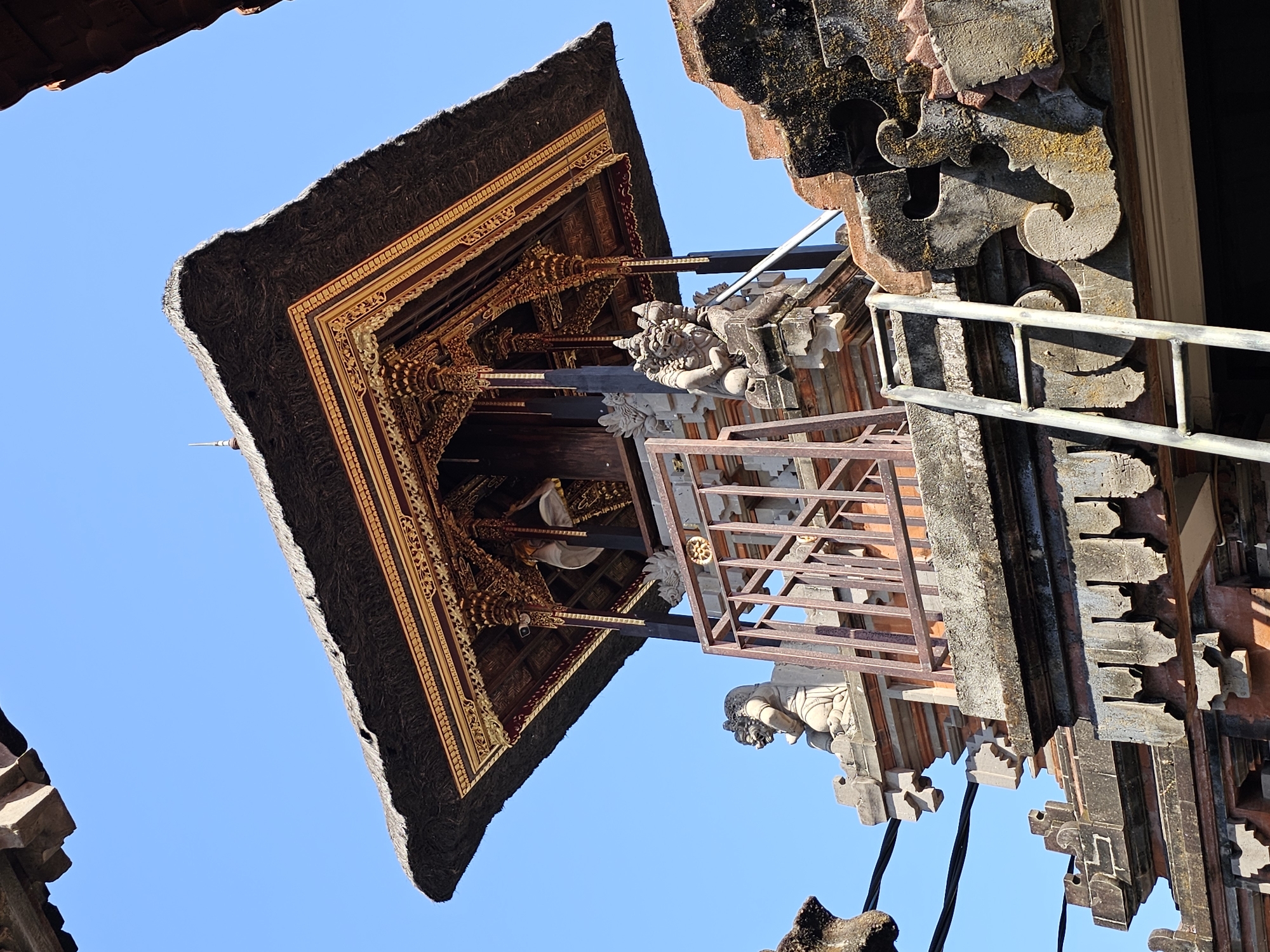 A traditional Balinese structure with a thatched roof, gilded trim, and stone carvings is depicted against a clear blue sky. The building features intricate details, including decorative pillars and what appear to be guardian statues on the rooftop. A metal ladder leans against the structure, suggesting ongoing maintenance or construction. Power lines are visible in the upper left corner. The architectural style and the clear blue sky indicate a location in a tropical or sub-tropical region, likely Denpasar, Indonesia, known for its elaborate temples and traditional buildings. The scene captures a moment of stillness, highlighting the craftsmanship and cultural heritage of the structure.