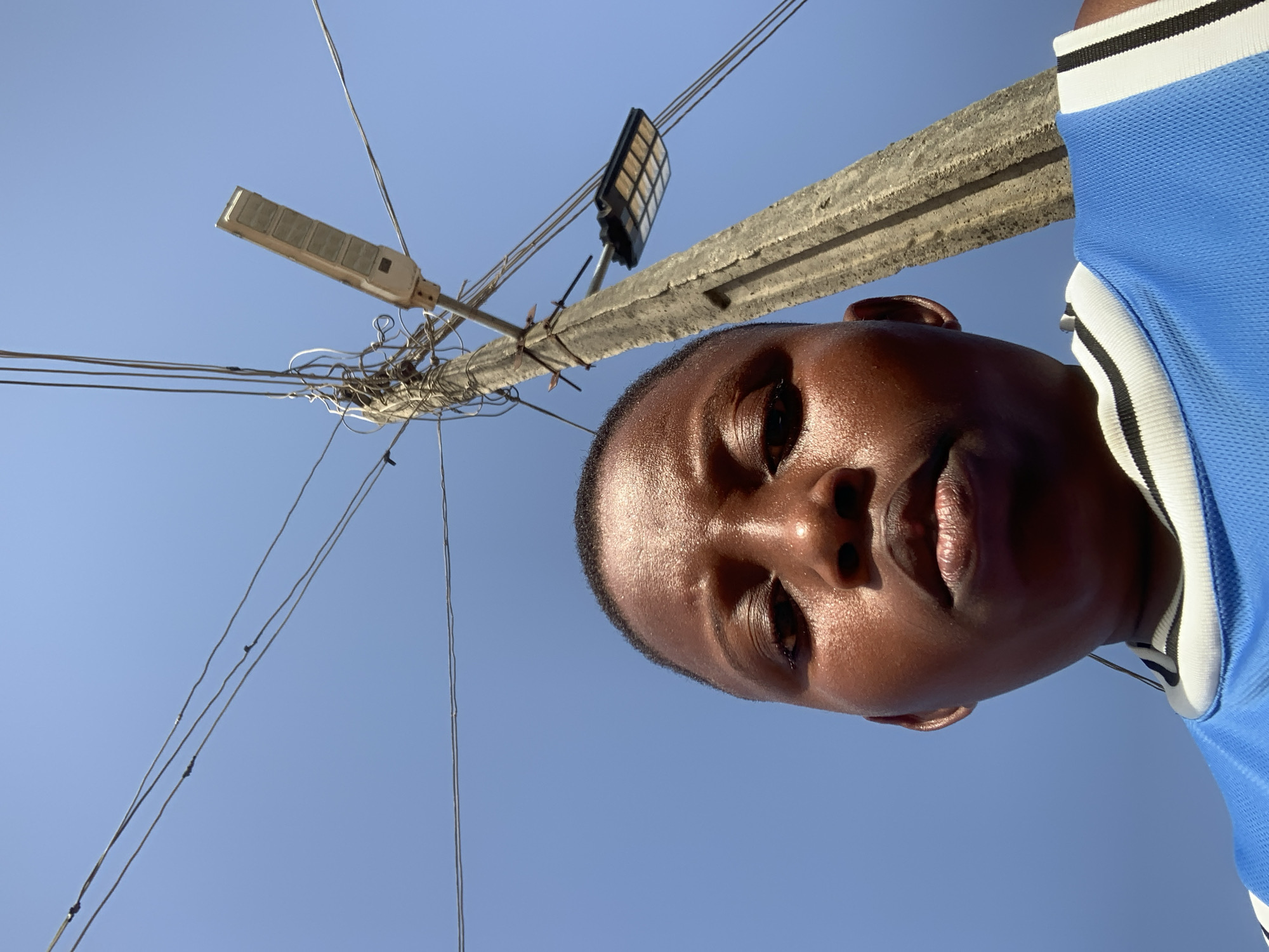 The image shows a low-angle view of a person with dark skin under a bright blue sky. The person is wearing a blue and white shirt. A concrete utility pole is visible, with a tangle of wires and two streetlights attached to it. One streetlight is a solar-powered model. The location is likely Ikorodu, Nigeria, based on the prompt.