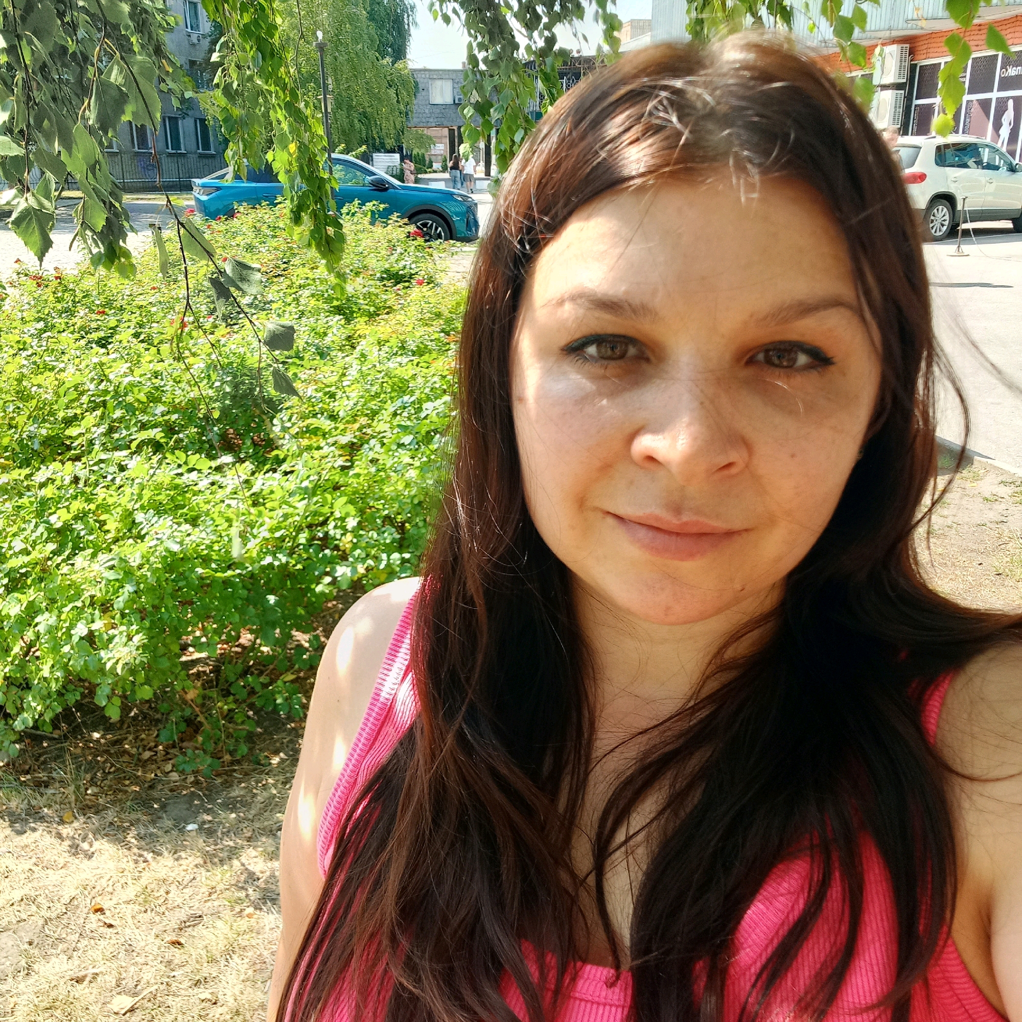 A woman with long, dark brown hair is taking a selfie outdoors in a city environment. She is wearing a bright pink, ribbed tank top. The background shows a green, leafy bush in the foreground and a street with buildings and vehicles behind it. A teal-colored car is parked to the left of the frame, and a light-colored SUV is visible on the right. The scene appears to be set in Kropyvnytskyi, Ukraine, as indicated by the presence of a building with Cyrillic signage that reads "OKSMO" on the right side of the image. The lighting suggests it is daytime, with sunlight casting shadows and highlights.