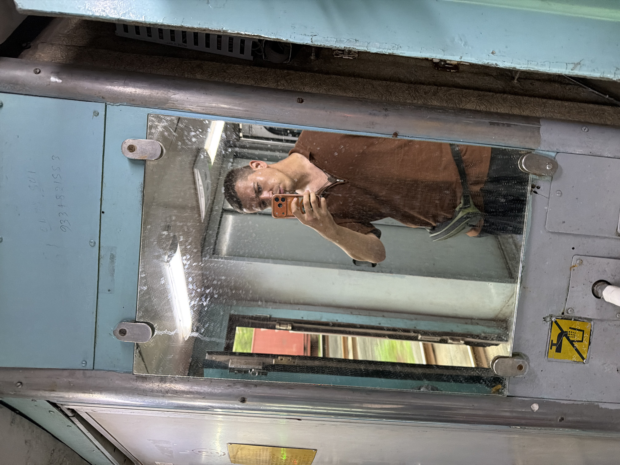 In Saphale, India, a man takes a mirror selfie inside what appears to be a train compartment. The man, wearing a brown polo shirt and dark pants, holds an orange smartphone to capture his reflection. He has short hair and a serious expression. The compartment features a large, grimy mirror that reflects him and the interior of the train. Visible in the reflection are fluorescent lights, metal fixtures, and a sliver of the outside world, showing greenery and train tracks through an open door. The overall environment is utilitarian, with faded blue and gray paint, metallic poles, and worn surfaces, indicating the age and use of the train car.