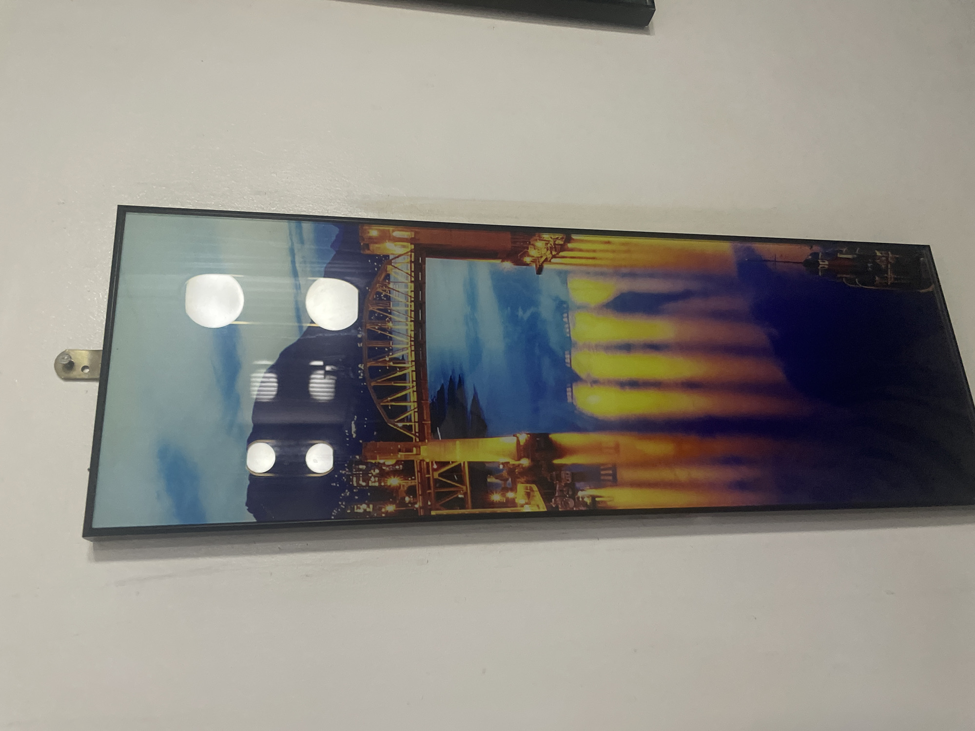 The image shows a framed, horizontal print hanging on a white wall. The print depicts a nighttime cityscape, likely reflecting on water. Key elements include a bridge with its structure lit up, and blurred orange and yellow streaks that likely represent the light trails of moving vehicles. The right side of the print is dominated by a darker blue or black area that may represent the night sky or dark water. Reflections are visible on the print's surface from overhead lights. The location cannot be determined from the image.