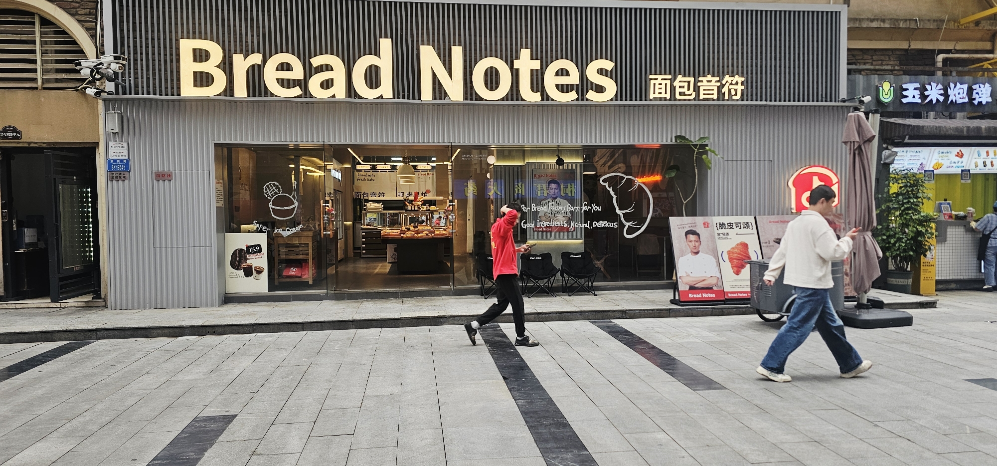 Here's a detailed description of the image:
**General Overview:**
The image captures a street scene in what appears to be a commercial area. The primary focus is on the front of a bakery called "Bread Notes" (面包音符). Two people are walking on the sidewalk in front of the bakery.
**Specific Details:**
* **Bakery "Bread Notes":** The bakery has a modern storefront with a gray façade and glass windows. A sign with the bakery's name in English and Chinese is prominently displayed. Inside, baked goods are visible. Promotional posters and displays are placed near the entrance, including images of chefs and croissants.
* **People:** There are two individuals walking on the sidewalk: one in a red sweatshirt and black pants, and the other in a light-colored jacket and blue jeans, pulling a cart.
* **Sidewalk & Street:** The sidewalk is paved with gray tiles and has dark stripes.
* **Surroundings:** To the left of the bakery is another building with a dark entrance. To the right is another establishment with a yellow sign with the characters 玉米炮弹. A tree in a planter is in front.
**Location Clues:**
* The Chinese signage and architecture suggest a location in China. The address in the upper left gives the number of the building 25, Unit 3 and the door number of 8954.
* The text from the OCR references 脆皮可颂 and 面包音符 which are the terms used to describe a bakery and a croissant.
* Given the details, this scene is located in Chongqing High-tech Zone, China.