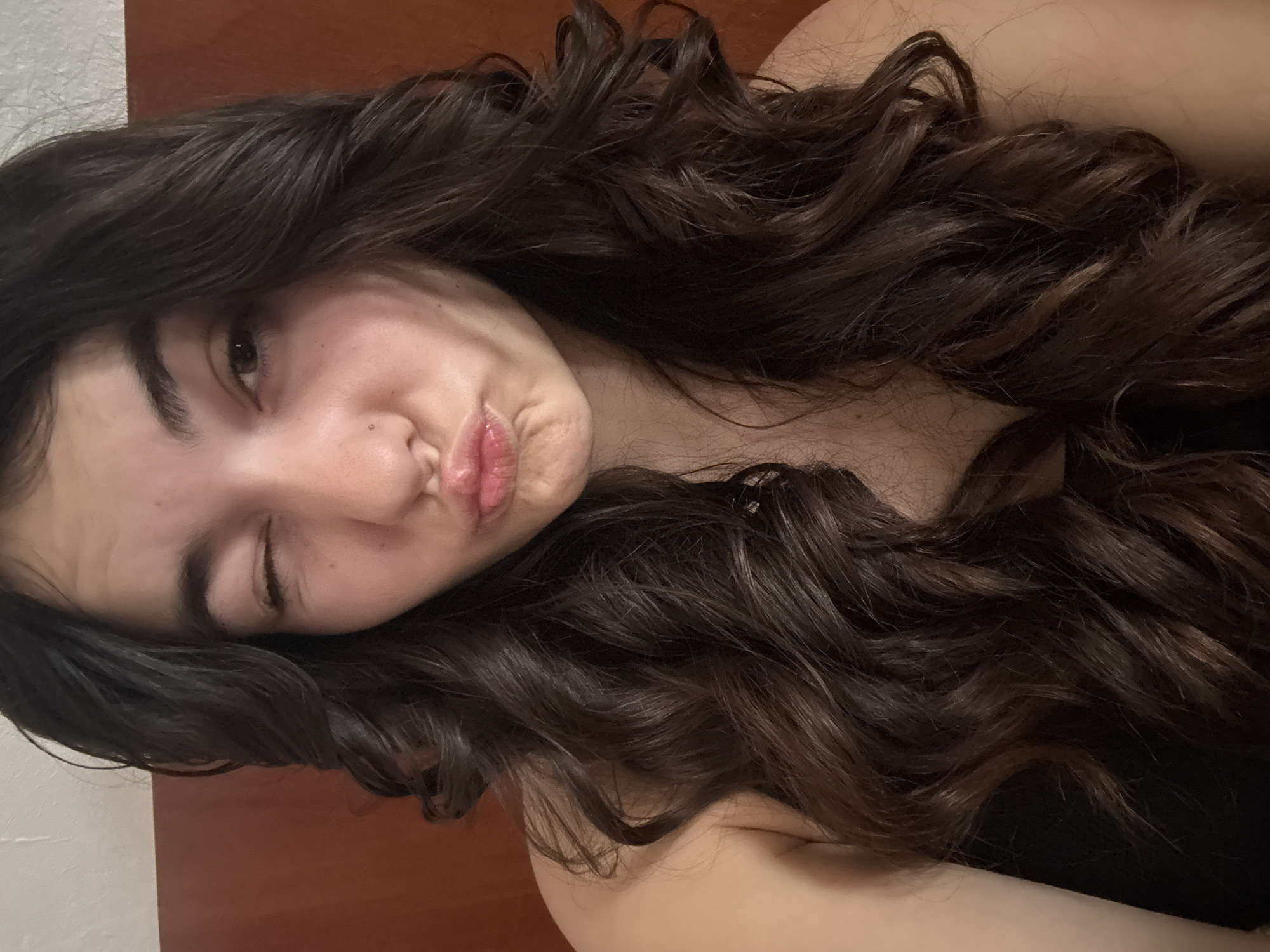 A brunette woman with long, wavy hair is making a kissing face and winking one eye. She appears to be lying down, with a dark garment visible below her neck. The background includes a wall that transitions from white to a darker shade, possibly wood paneling. The location cannot be determined to be in the city of Plovdiv, Bulgaria from the image.