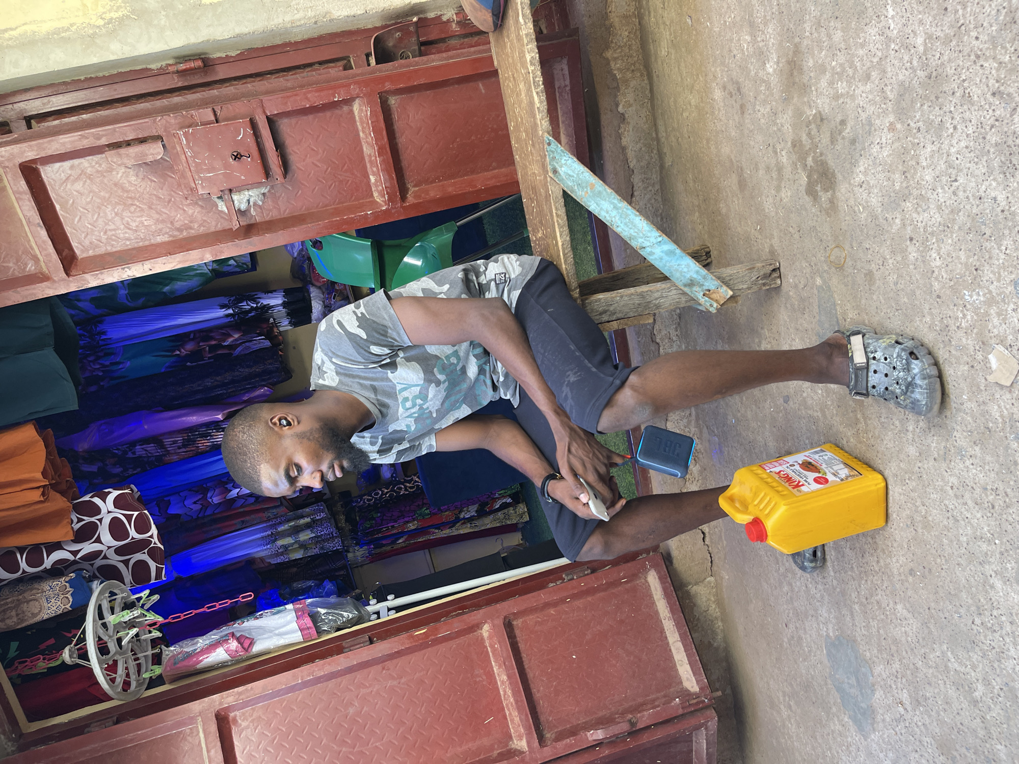 A young adult male, dressed in a grey camouflage t-shirt and black shorts, sits on a makeshift wooden bench in front of a market stall or small shop in Samaru, Nigeria. His right foot wears a grey, open-toed sandal, while his left foot is bare. He is engrossed in a white smartphone held in his hands.
Beside him, on the rough concrete ground, a small black JBL portable speaker is visible. To his left, a bright yellow plastic container with a red cap, labeled "KING'S VEGETABLE OIL," rests on the ground. The background features reddish-brown metal bi-fold doors with a diamond plate texture, which form the shop's entrance. Inside the shop, various colorful fabrics, garments, and what appears to be an umbrella are displayed on racks and shelves, indicating a retail business. A circular clothes hanger with pegs is also visible.
The scene depicts a typical daytime setting, likely a street-side shop or market stall, with the proprietor attending to his business while utilizing his phone during a moment of calm.