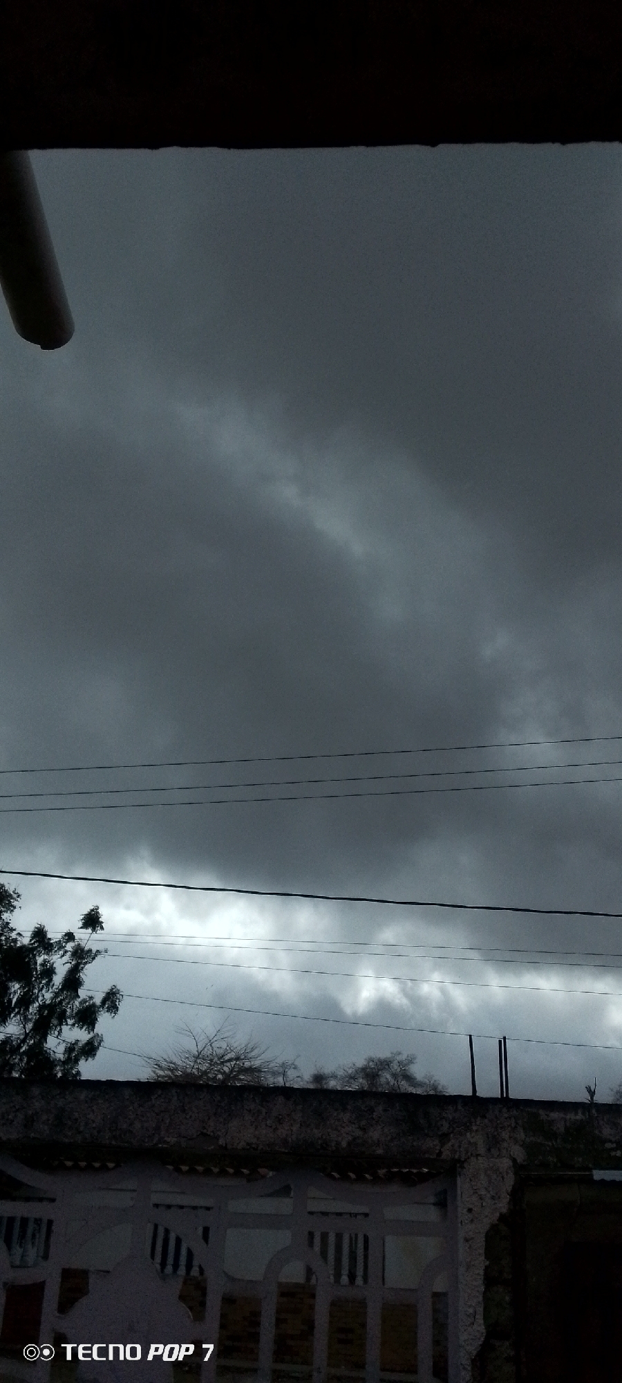 The image captures a somber, heavily overcast sky, likely indicating stormy weather or an approaching downpour. Dark, dense grey clouds fill the majority of the frame, with some lighter grey patches visible, particularly in the middle and lower sky sections.
At the very top, a dark, solid overhead structure, possibly part of a building's roof or an overhang, is visible, partially obscuring the uppermost clouds. A dark, pipe-like object hangs from this structure on the upper left. Multiple thin, dark power lines or cables run horizontally across the mid-section of the sky, segmenting the cloud patterns.
Below the horizon line created by the lower clouds, the upper parts of trees are visible. On the left, a tree with dark, sparse foliage is partially seen, while on the right, what appear to be bare branches extend above a wall. The foreground features a white or light-colored, patterned fence or wall with distinct horizontal bars and arched details, topped by a textured, darker coping. Behind this barrier, dimly visible structures suggest a residential or built-up area in Altagracia, Venezuela.
The overall impression is one of low light and impending weather, suggesting either an early morning, late afternoon, or a dark day due to the cloud cover. There is no visible human activity. In the bottom left corner, a "TECNO POP 7" watermark is present.