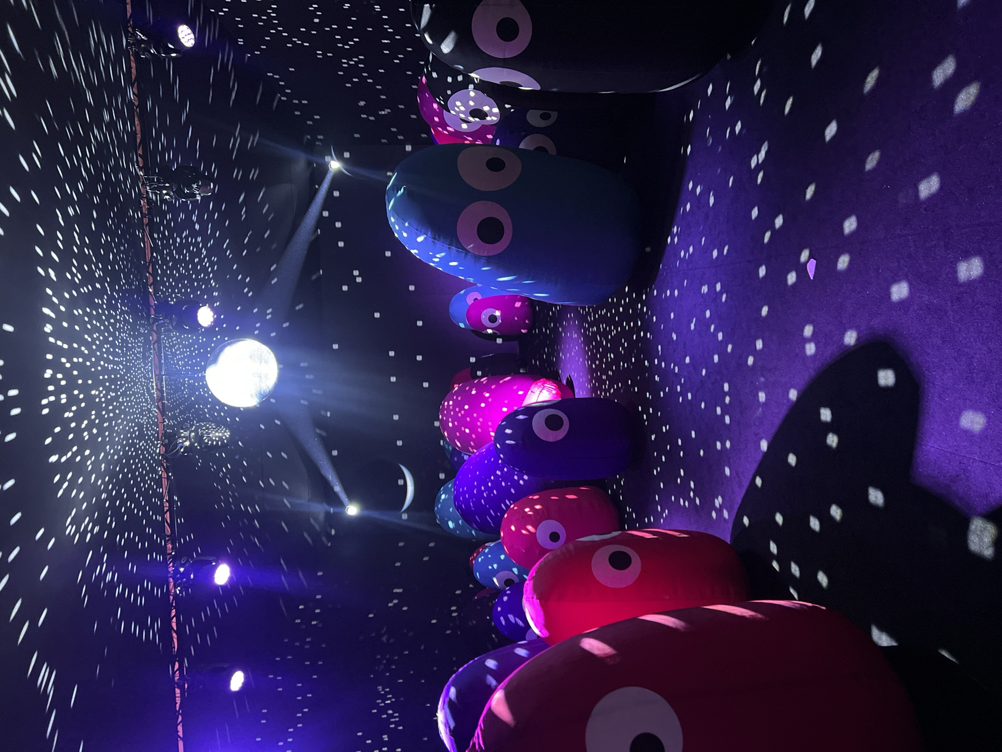 An immersive art installation is depicted in this image, likely a nighttime or indoor event in Miami, USA. The scene is characterized by a dark, cavernous space illuminated by various light sources and projections. The ceiling is adorned with a dense pattern of small, white lights, resembling stars, which are also projected onto the floor, creating a dazzling, speckled effect. A prominent disco ball hangs from the ceiling, catching and reflecting light. Several spotlights are visible, casting beams of purple and white light across the room.
The primary subjects are a collection of large, inflatable, bean-shaped figures with simple, cartoonish faces, featuring large circular eyes with black pupils and pink irises. These figures are arranged throughout the space in varying sizes and colors, including bright red, pink, blue, purple, and black. Some are clustered together, while others stand more isolated. The floor is a dark purple carpet, also illuminated by the projected white light patterns. The overall atmosphere is whimsical and surreal, designed to evoke a sense of wonder and playfulness, fitting for an art exhibit or interactive entertainment venue in Miami.