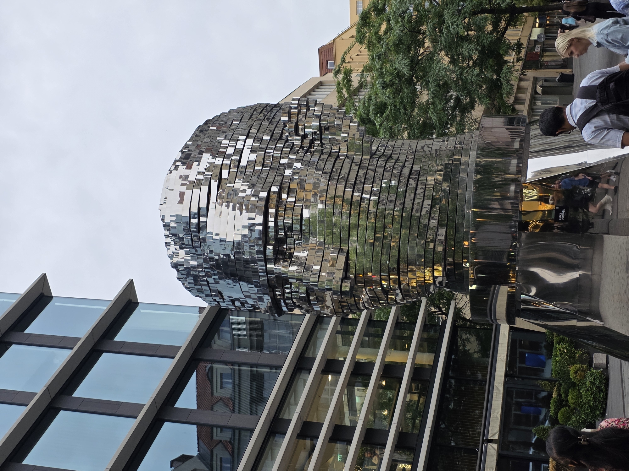 A large, multi-layered, reflective metallic sculpture of a human head dominates the center-right of the frame. The head is constructed from numerous horizontal, segmented plates that create a dynamic, fragmented appearance.
On the right side of the image, several people are present: a blonde-haired individual in a light top and another with dark hair wearing a light shirt and a backpack, both looking downwards, possibly observing the sculpture's base or the ground. Other indistinct figures appear in the background and as reflections on the sculpture's surface.
To the left of the sculpture is a modern building with large glass windows and dark frames, reflecting parts of the urban surroundings. Older buildings with lighter facades and green trees are visible in the background, particularly in the upper right. Green foliage, including trees and shrubs, is also present around the sculpture's base, providing a natural contrast to the metallic structure.
The highly polished, chrome-like surface of the sculpture vividly reflects the urban environment, including buildings, trees, people, and the overcast sky, enhancing its complex visual effect. Faint white text, "KAFKA PORTAL," is visible in one of the reflections near the base of the sculpture. The scene depicts a public art installation in an urban setting during what appears to be an overcast daytime.