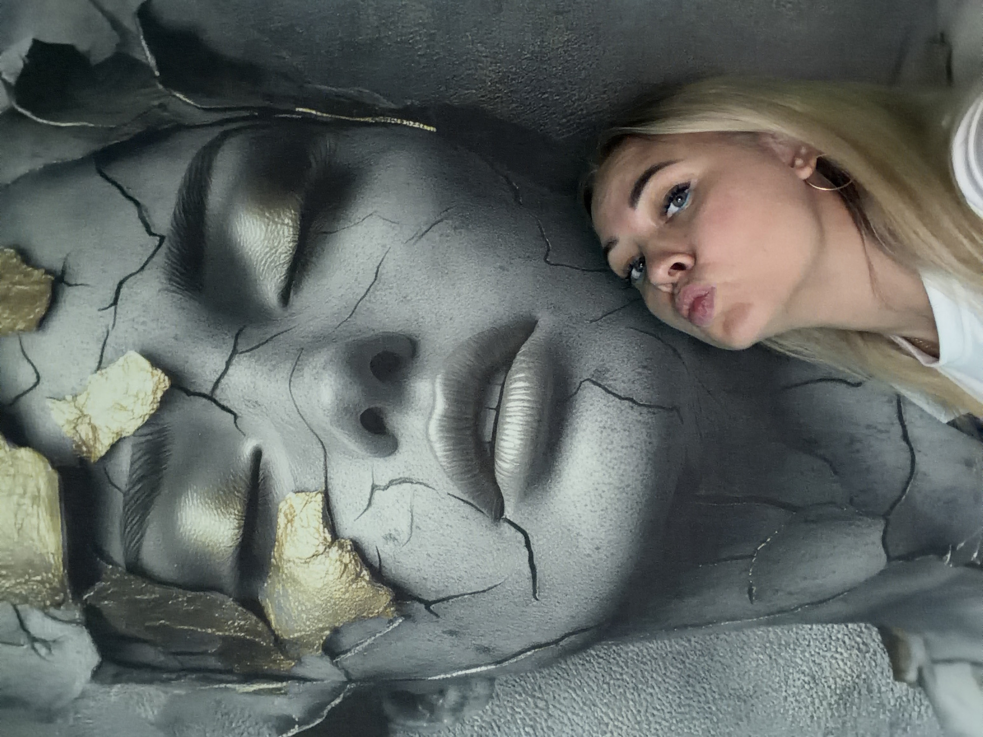 In this media file, a young woman with blonde hair and blue eyes leans in towards a large, cracked sculpture of a human face. The sculpture is grey and appears to be made of stone or a similar material, with gold leaf accents scattered across its surface, particularly on the cheeks and forehead. The woman is puckering her lips, as if blowing a kiss or making a funny face for the camera. She is wearing a white t-shirt and a hoop earring in her right ear. The background is dark and textured, suggesting an indoor or dimly lit environment. The overall impression is playful and artistic, with the woman interacting with the striking sculpture. The location is Chaikovsky, Russia. There is no visible text in the image.
