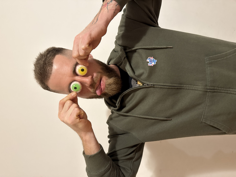 A man poses playfully, holding two candy eyeballs in front of his face. One eyeball is yellow, and the other is green. He wears a dark green zip-up hoodie with a cartoon character pin on the left breast. The man has a beard and short hair and is sticking his tongue out. Tattoos are visible on his left forearm. The background is a plain, light-colored wall. The image is slightly rotated.