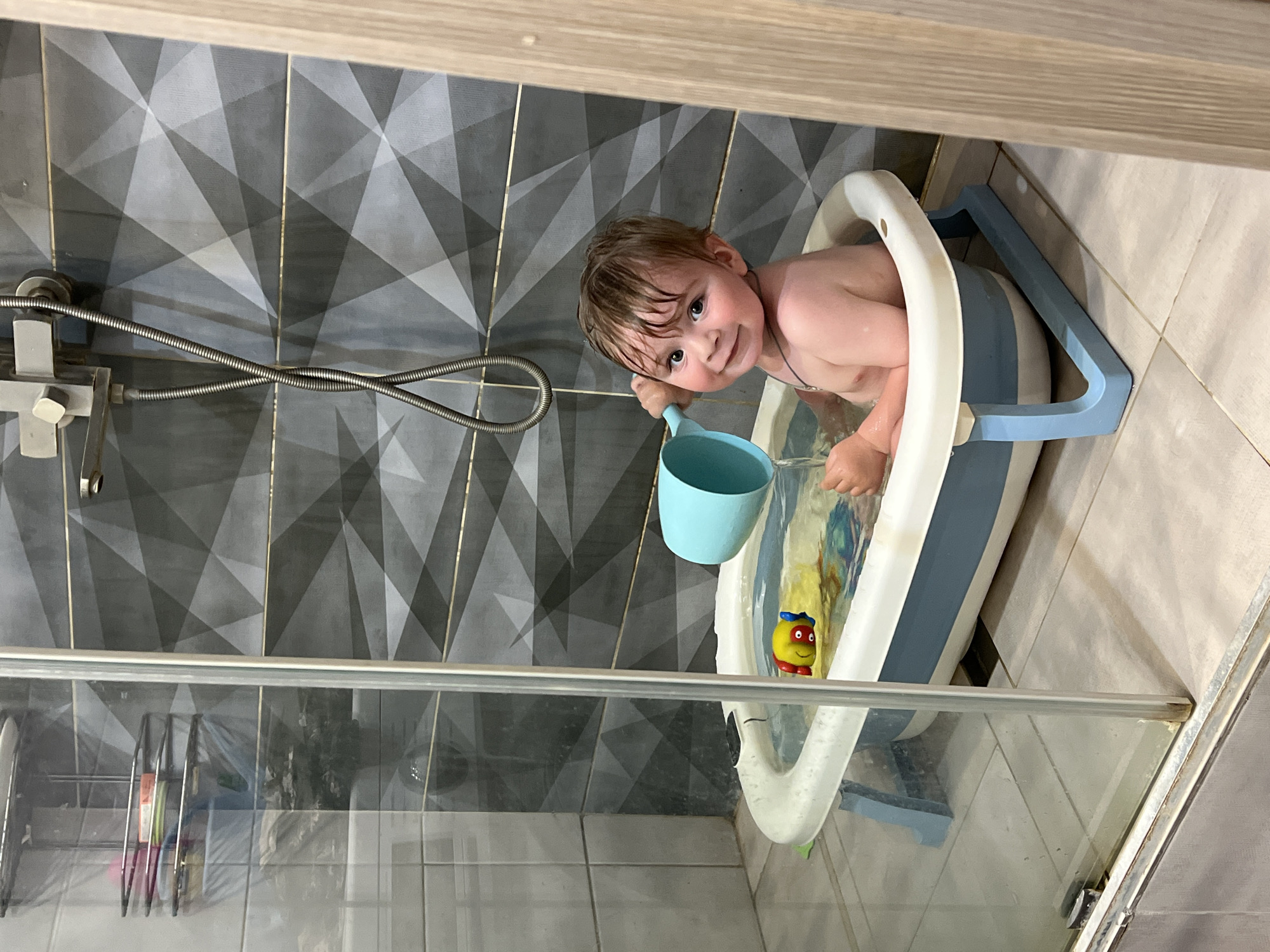 A young child is sitting in a small bathtub filled with water, located on the floor of a bathroom in Noviy, Russia. The child, with wet hair and a happy expression, is holding a blue watering can and appears to be pouring water. A yellow toy fish is floating in the tub. The bathroom features grey patterned tiles on the wall behind the tub, a silver shower head and hose, and a glass shower enclosure. A shelf with toiletries is visible through the glass.