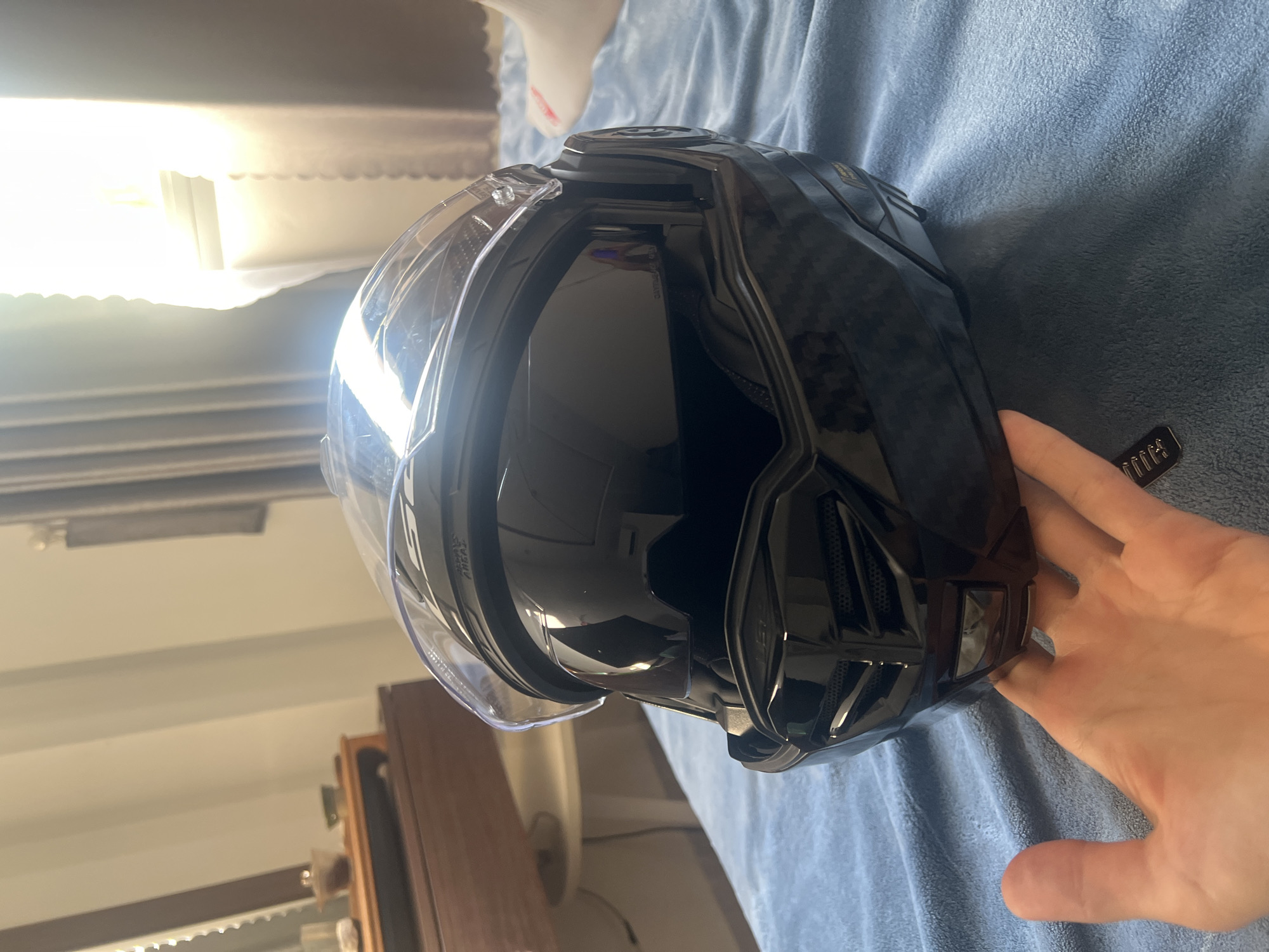 An overhead, slightly angled, medium shot captures a black motorcycle helmet resting on a plush, blue blanket. The helmet, with its visor partially open, displays a dark, glossy finish that reflects the bright sunlight streaming in from a window. The light illuminates the textured surface of the helmet and casts a strong glare across the visor. In the foreground, a human hand with visible palm lines is positioned to support the helmet from underneath. The background suggests an indoor setting, possibly a bedroom, with hints of furniture and curtains. The image does not provide specific geographical indicators for Đà Nẵng, Vietnam.