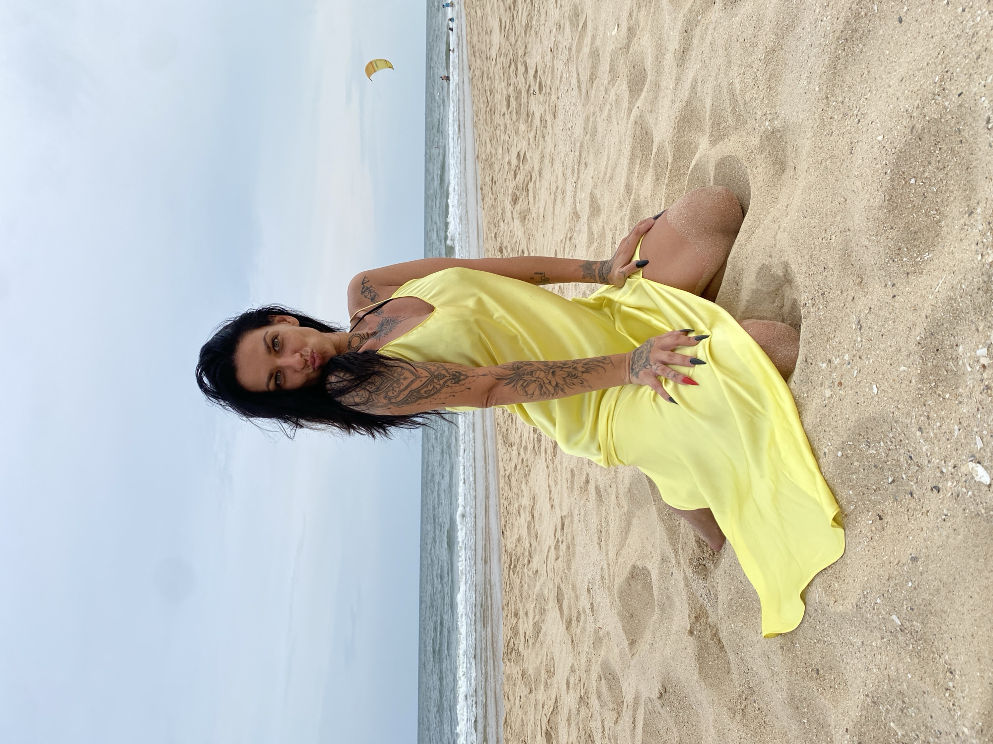 A woman with dark hair and tattoos is kneeling on a sandy beach in Phu Thuy Ward, Vietnam. She is wearing a yellow dress. The ocean is in the background with waves breaking on the shore. A kite can be seen flying in the sky. The sky is overcast.