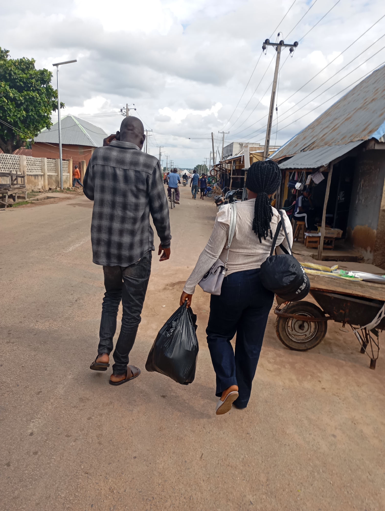 A man and a woman are walking away from the camera on a dirt road in Shendam, Nigeria. The man is wearing a grey and black plaid shirt and dark jeans. He is talking on his phone and wearing brown sandals. The woman is wearing a cream-colored long-sleeved shirt and dark blue jeans. She is carrying a black plastic bag in her right hand and has a black duffel bag slung over her left shoulder. Her left hand is resting on the strap of a grey handbag. In the background, there are buildings with corrugated metal roofs, utility poles with wires, and a few other people. A cart with a wheel is visible to the right of the woman. The sky is cloudy.