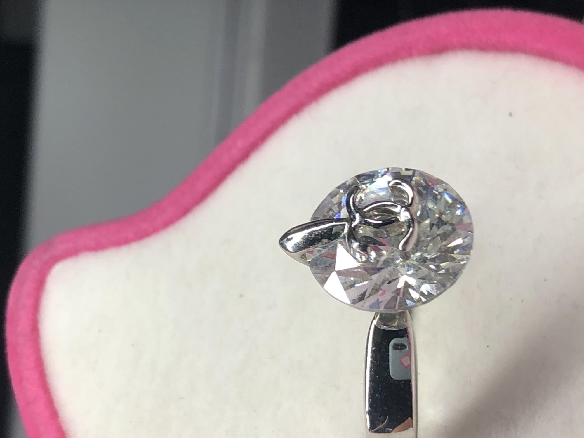 A close-up, eye-level, indoor shot shows a sparkly, faceted gemstone with a small, silver charm attached to its top. The gemstone is round and appears to be a diamond, with a setting at the top that allows it to be attached to a chain or other jewelry. The charm is a stylized "CC" logo, likely representing a brand. The gemstone is positioned on a white, fuzzy surface, bordered by a bright pink fuzzy material on the right and top. To the left of the gemstone, a silver object, possibly a pair of tweezers or a jewelry clamp, is visible, with a reflection of a pink object on its surface. The background is out of focus, with dark and gray tones. There is no indication of the city of Yelizovo, Russia, or any outdoor elements in the image.