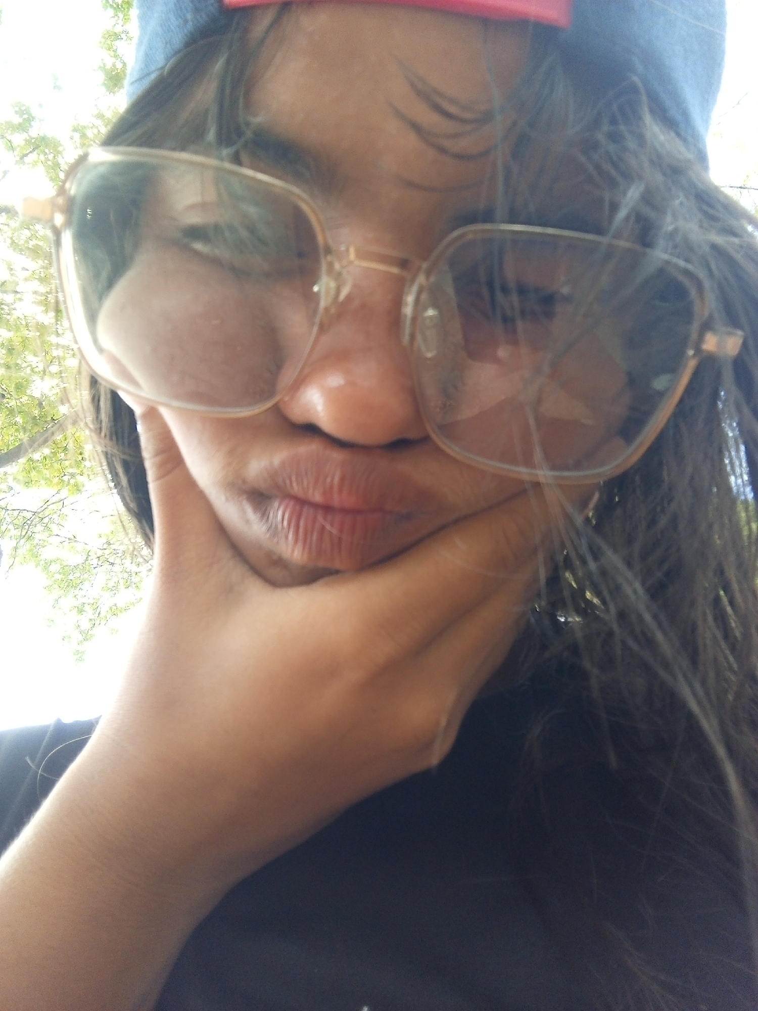 A selfie of a person with long, dark, slightly disheveled hair wearing large, square-framed glasses and a blue cap. The person's face, partially obscured by the glasses, is pressed against their hand, creating a pouting expression. The person is wearing a dark-colored shirt. The background is blurry and appears to be foliage, indicating an outdoor location. No specific details suggest the image was taken in Juan Griego, Venezuela.