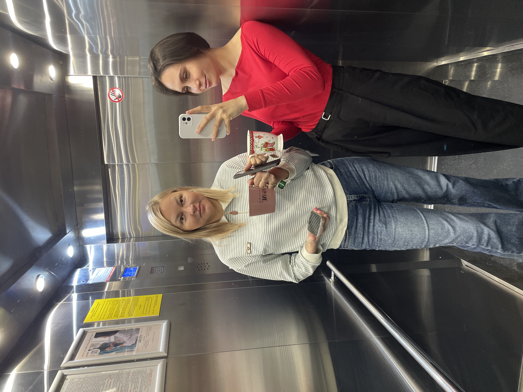 Two women are in an elevator with stainless steel walls. The woman on the top is wearing a red long-sleeved shirt and black pants and is holding a white smartphone and a Christmas-themed mug. The woman on the bottom is wearing a striped hoodie and jeans and is holding a black smartphone and a passport. They are taking a selfie. There is an out-of-focus notice in the elevator with text visible on it. Based on text visible on the notice, the location is Izhevsk, Russia.