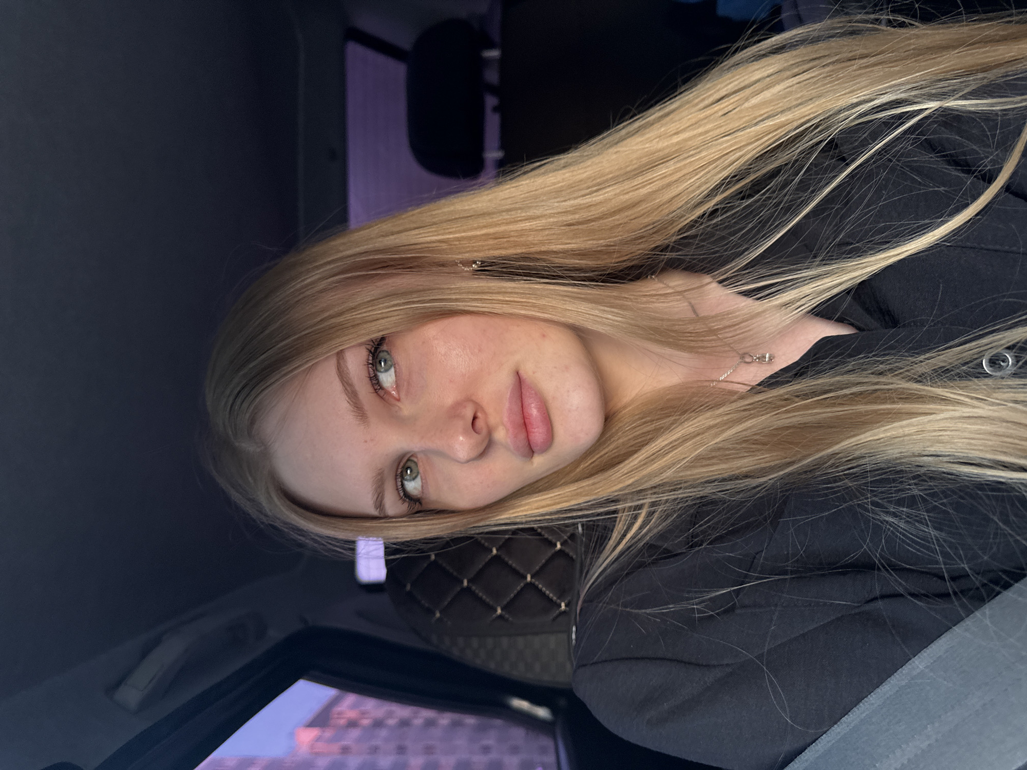 A young woman with long, straight, blonde hair is sitting in a car. She is wearing a black hooded sweatshirt. The background suggests an urban environment with buildings visible through the car window. The location may be Кондратово, Russia. The scene is captured in a close-up, portrait-style composition.