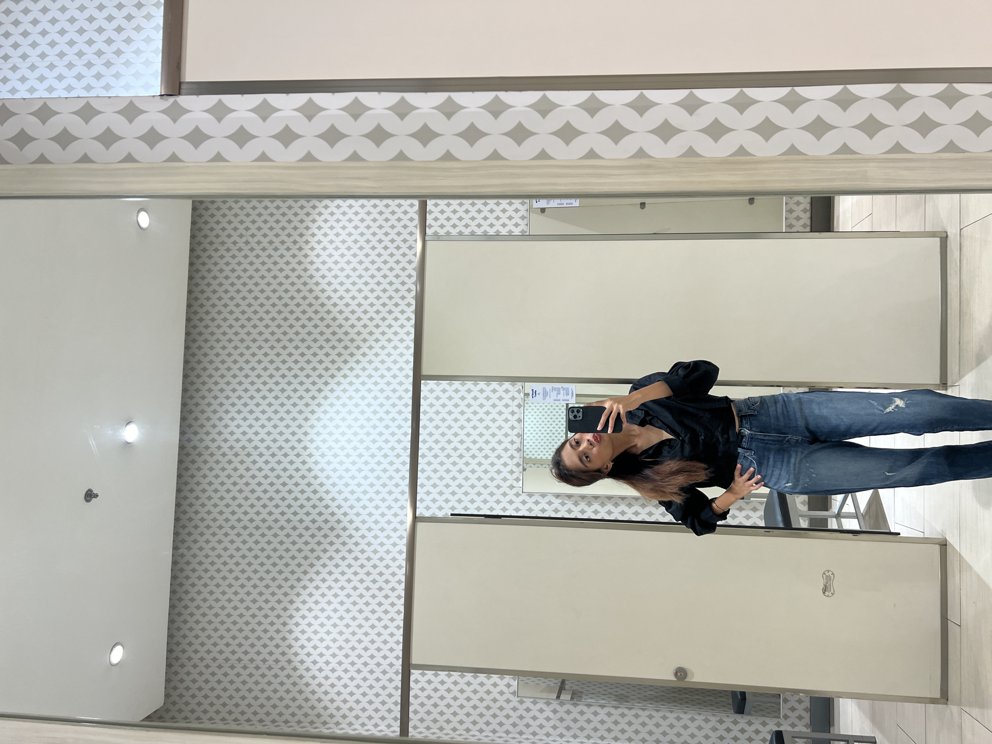 A young female is depicted in a mirror selfie within a fitting room, likely in a retail establishment in Davao City, Philippines. She has medium-brown hair, dark eyes, and is posing with puckered lips while holding a black smartphone with a black case to capture the image. She is wearing a black long-sleeved cropped collared shirt and distressed blue jeans. Her right hand is placed on her hip. The fitting room features light grey wallpaper adorned with a repeating white geometric pattern of interlocking circles. The ceiling is white, with two recessed LED spotlights and a circular ceiling fixture (possibly a smoke detector or sprinkler head) visible in the reflection. The floor is covered with light-colored tiles. Two cream-colored fitting room doors are visible in the background reflection; one on the left is mostly closed with a metallic handle, and one on the right is slightly ajar, revealing a dark stool or bench inside. Vertical wooden trim with the same patterned wallpaper frames the main mirror. A small, oval-shaped metallic sign is affixed to the lower section of the left door. A smaller mirror or reflective surface visible in the phone's reflection shows text, partially legible as "at least 1 type."