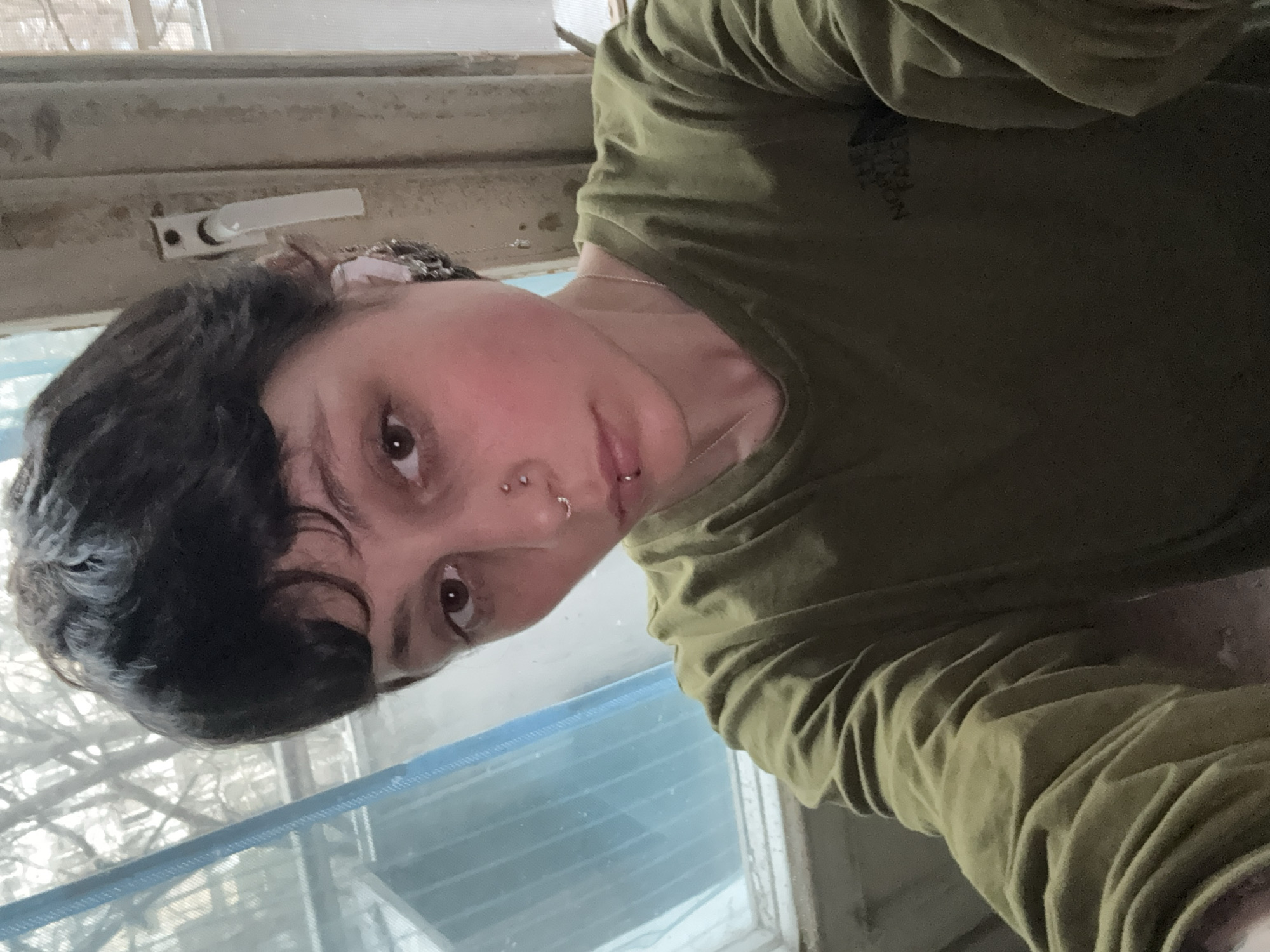 A person with dark hair and fair skin is wearing a green shirt. They have multiple piercings in their nose and lip. The background shows a window with a screen. The window frame appears weathered and shows traces of a building outside. There's no evidence to determine the location as Moscow, Russia.