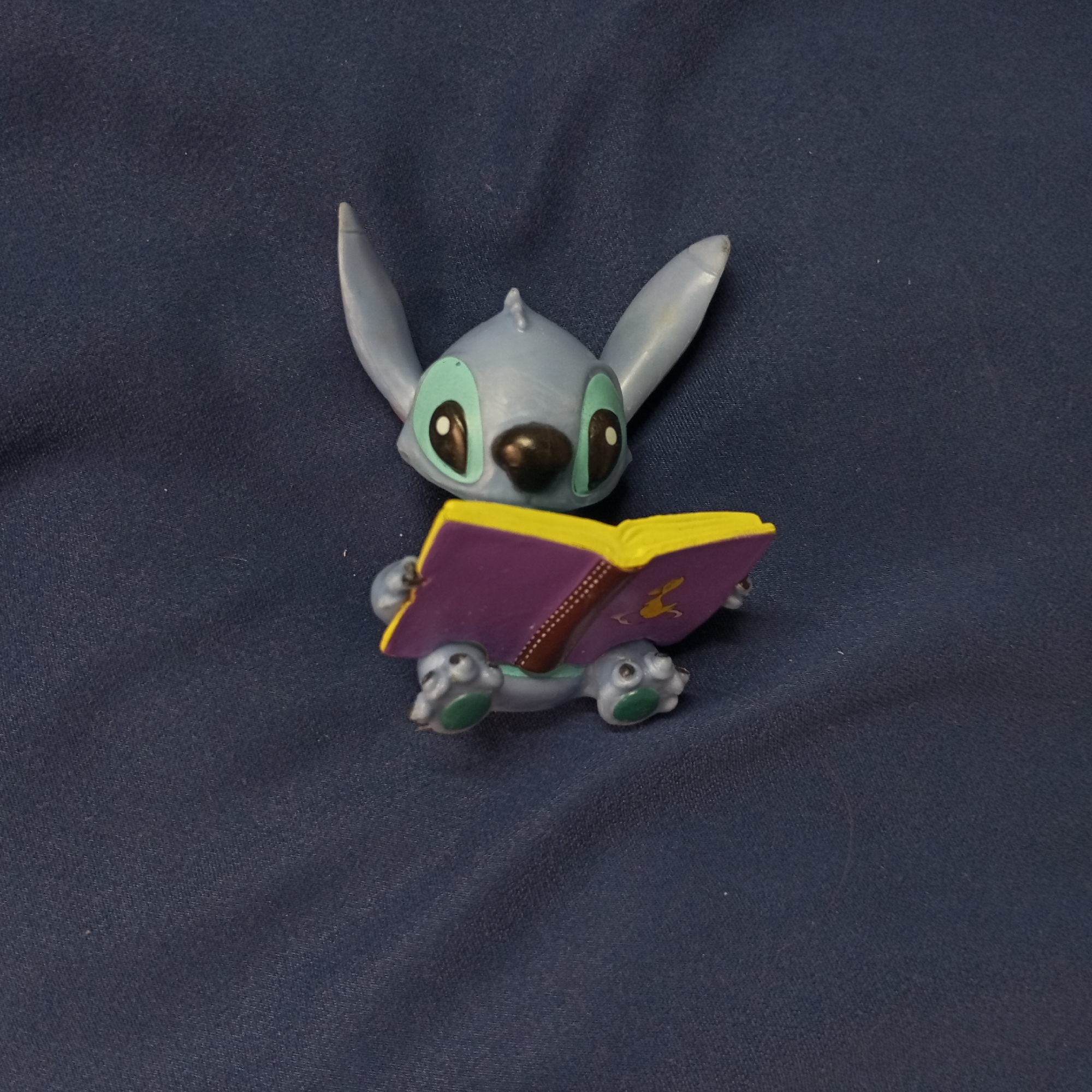 A figurine of Stitch from Disney's "Lilo & Stitch" is positioned on a dark blue fabric surface. The figurine is a light blue and depicts Stitch sitting while holding a book with a purple cover, yellow pages, and a brown spine. A stylized gold image is on the book cover. Stitch's feet are visible, with green paw pads. The background is a plain, dark blue fabric texture. The scene is indoors. The city of Pervomaysky, Russia is not indicated in the image.