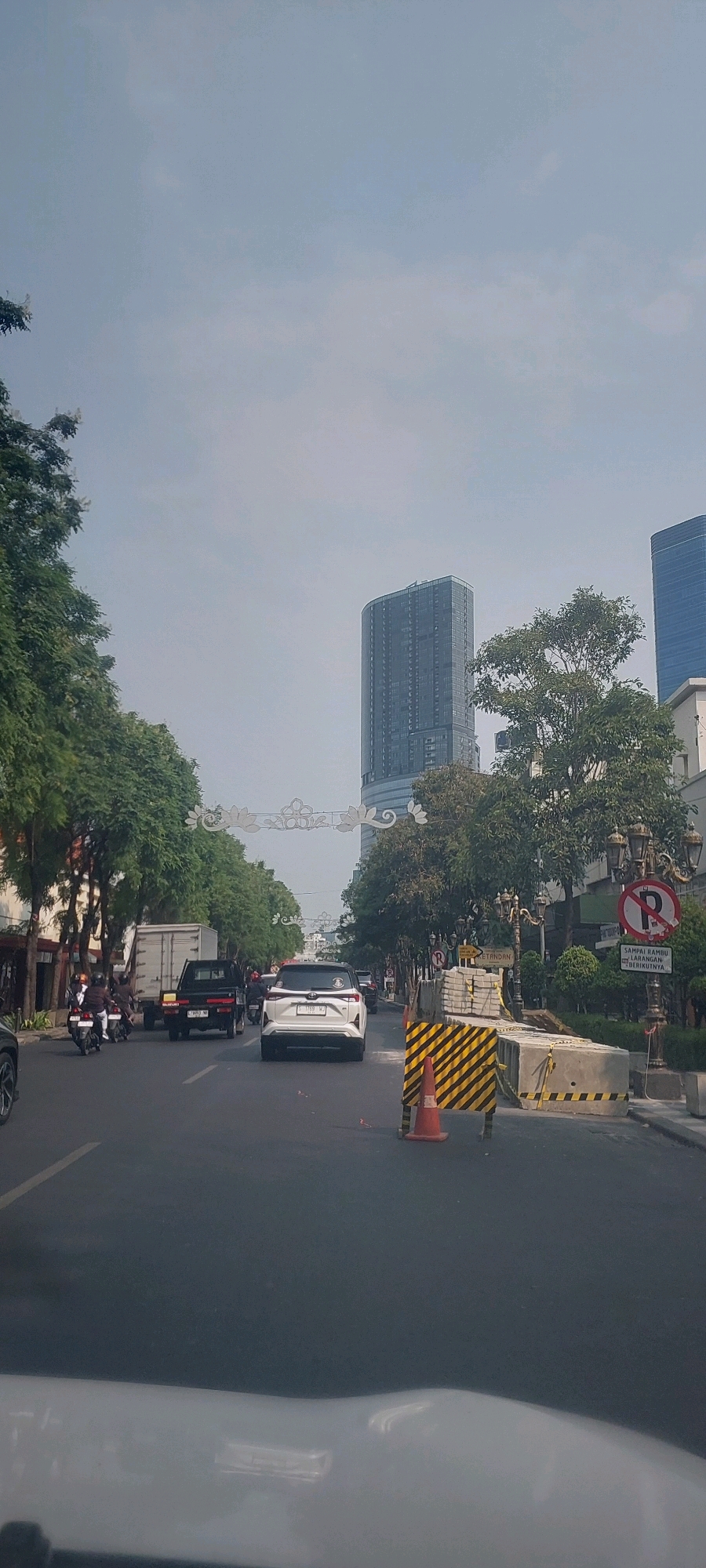 The image depicts a street scene in Surabaya, Indonesia, likely during daytime under an overcast sky. The perspective is from inside a vehicle, looking forward. On the right side of the road, there are construction barriers, a traffic cone, and a lamppost with a "No Parking" sign (a red circle with a diagonal line and a 'P'). Further back, trees and modern buildings, including a tall skyscraper, are visible. On the left side of the road, there are more trees, a white truck, a black pickup truck, and several motorcycles with riders. The road is asphalt and marked with white lines. A white car is in the foreground, facing away from the viewer. There are decorative white archways strung across the street in the middle distance.
