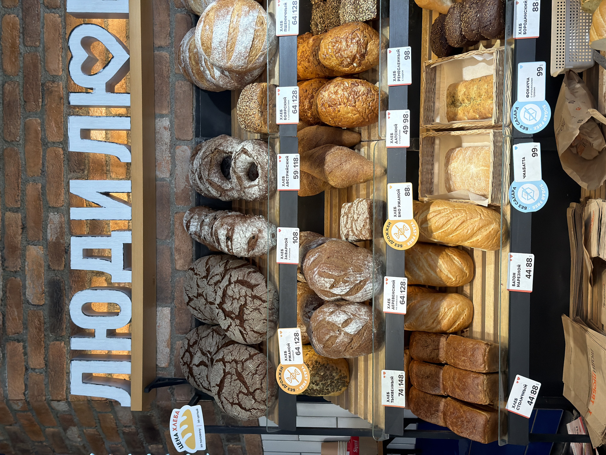 This is an eye-level, indoor shot of a bakery display, most likely located in Saint Petersburg, Russia. The display features a variety of breads arranged on shelves, with each type labeled with its name and price in Russian currency. The breads include rye, sourdough, pumpkin, and ciabatta varieties, among others. Some are identified as "sugar-free" and "yeast-free." There is a decorative sign with the word "ЛЮДИ ЛЮ" in Cyrillic script, which translates to "People Love" in English. The bakery’s name is in vertical, white block letters, with the heart shape replacing the "O" in "love," set against a brick wall backdrop. Prices and bread names are listed next to each product. Some of the items have "sugar-free" labels. The overall impression is one of a well-stocked bakery offering a diverse selection of bread.