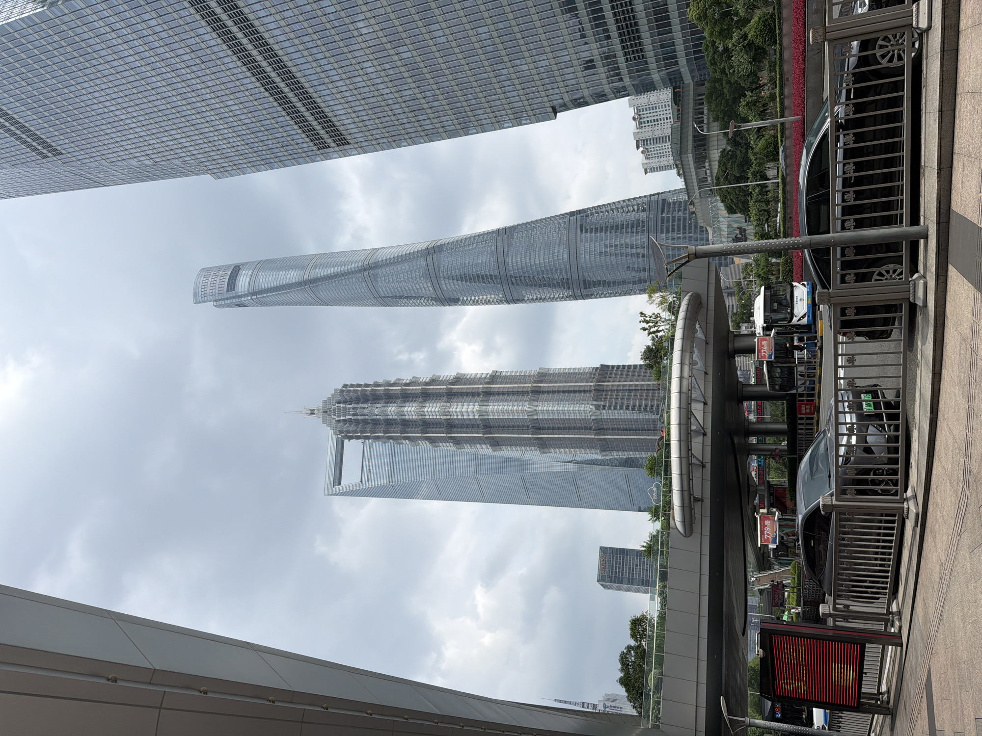 The image depicts a street-level view of Pudong, China, with a focus on the iconic Shanghai World Financial Center, Jin Mao Tower, and Shanghai Tower. The foreground shows a sidewalk with a metal railing, and a dark-colored car is parked behind it. A red digital sign is visible to the left. Above the street level, an elevated pedestrian walkway or overpass is present, with buses and other vehicles passing underneath. The background is dominated by the impressive skyscrapers, characterized by their modern architectural designs. The sky is overcast, indicating a cloudy day. Lush greenery and some smaller buildings are interspersed between the massive structures, adding a touch of nature to the urban landscape.