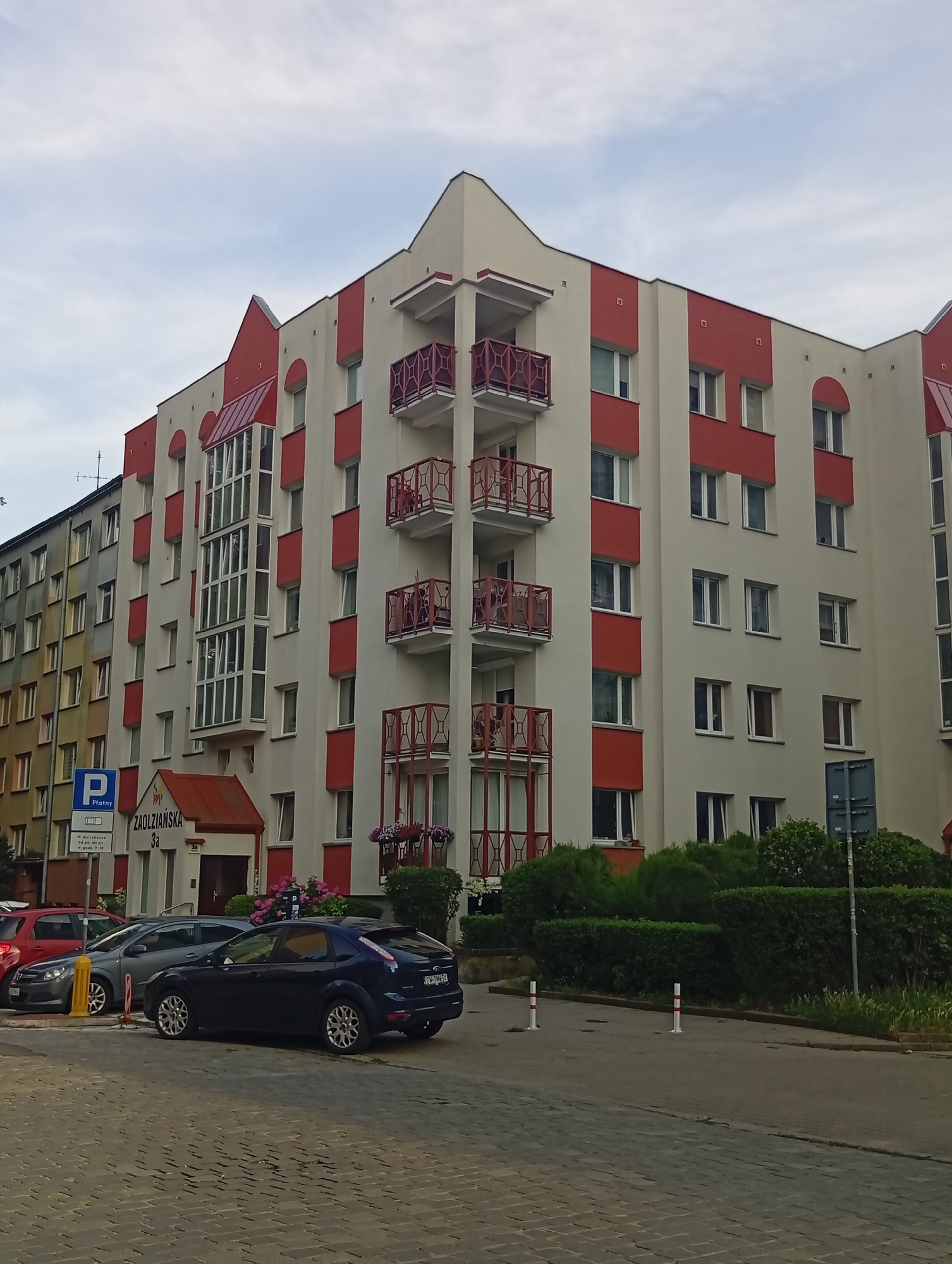 The image depicts a modern apartment building on Zaolziańska Street 3a in Wrocław, Poland. The building features a cream-colored facade accented with red panels and decorative balconies with red railings. A glass-enclosed section with multiple windows is visible on the left side of the building. A red awning extends over the entrance. In front of the building, several cars are parked, including a dark blue Ford Focus. A blue and white parking sign with Polish text is visible on the left side of the image. The street in the foreground is paved with cobblestones. Green bushes and hedges line the front of the building and the street. The sky is overcast.