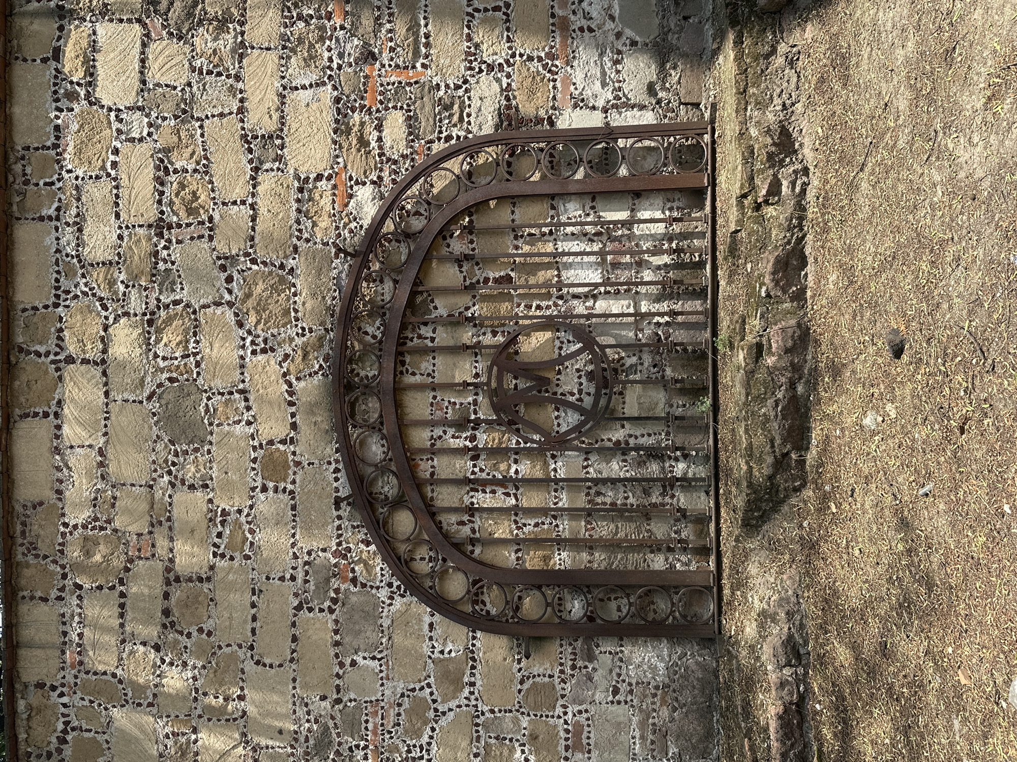 The image captures a weathered stone wall in Tlalnepantla, Mexico. The wall's construction consists of rectangular stones arranged in a grid pattern with smaller dark stones filling the gaps. An ornate, rusted metal gate is affixed to the wall, its design featuring a series of circles along the top arch and horizontal bars in the main section. A circular emblem is located in the center of the gate. To the right of the wall is a section of rough, earthen terrain covered in dry vegetation. The lighting is bright, casting shadows that accentuate the textures of the wall and the gate.